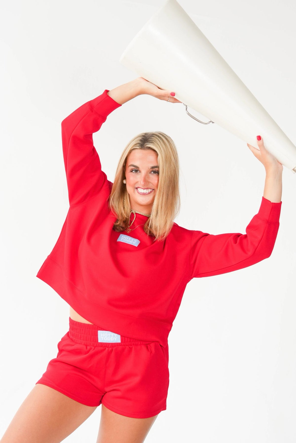 A smiling blonde woman in a bright red cropped long-sleeve top and matching red shorts holds a large white megaphone against a plain white studio background.