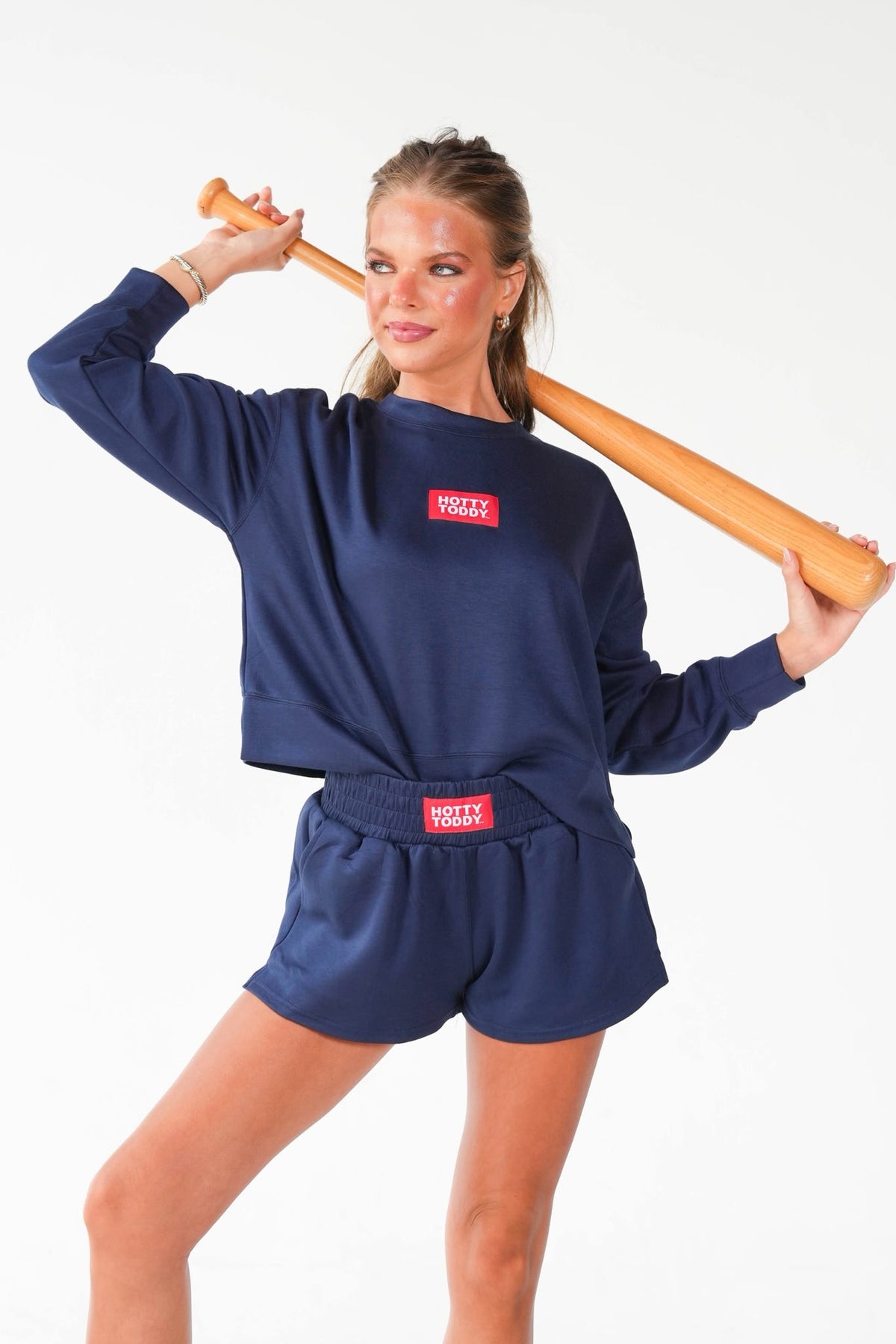A woman modeling a navy blue cropped sweatshirt and matching shorts set with a red rectangular chest patch that reads 'VOGUE'. She is holding a wooden baseball bat over her shoulder against a plain white studio background.