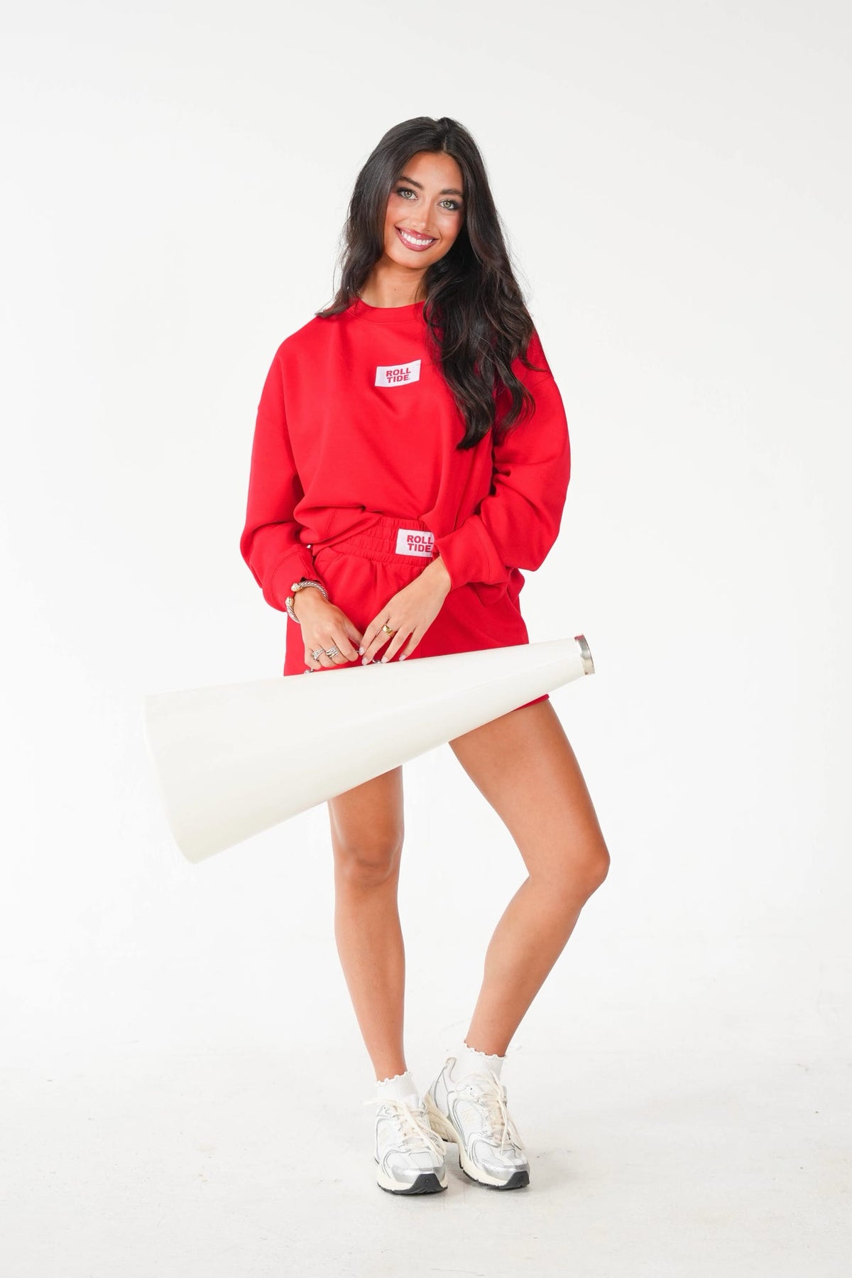 A woman modeling a bright red sweatshirt and matching shorts set, holding a large white megaphone prop against a seamless white studio background.