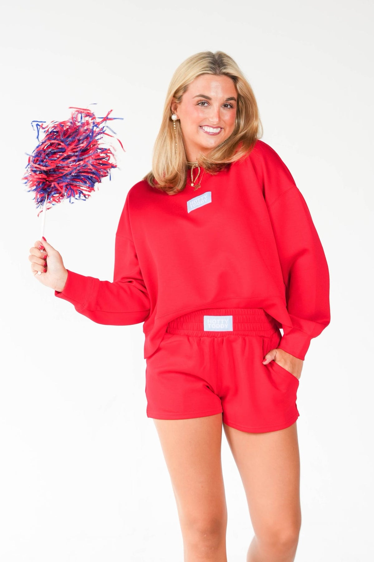 A woman modeling a bright red sweatshirt and matching shorts set, holding a small red and blue pom-pom against a plain white background.