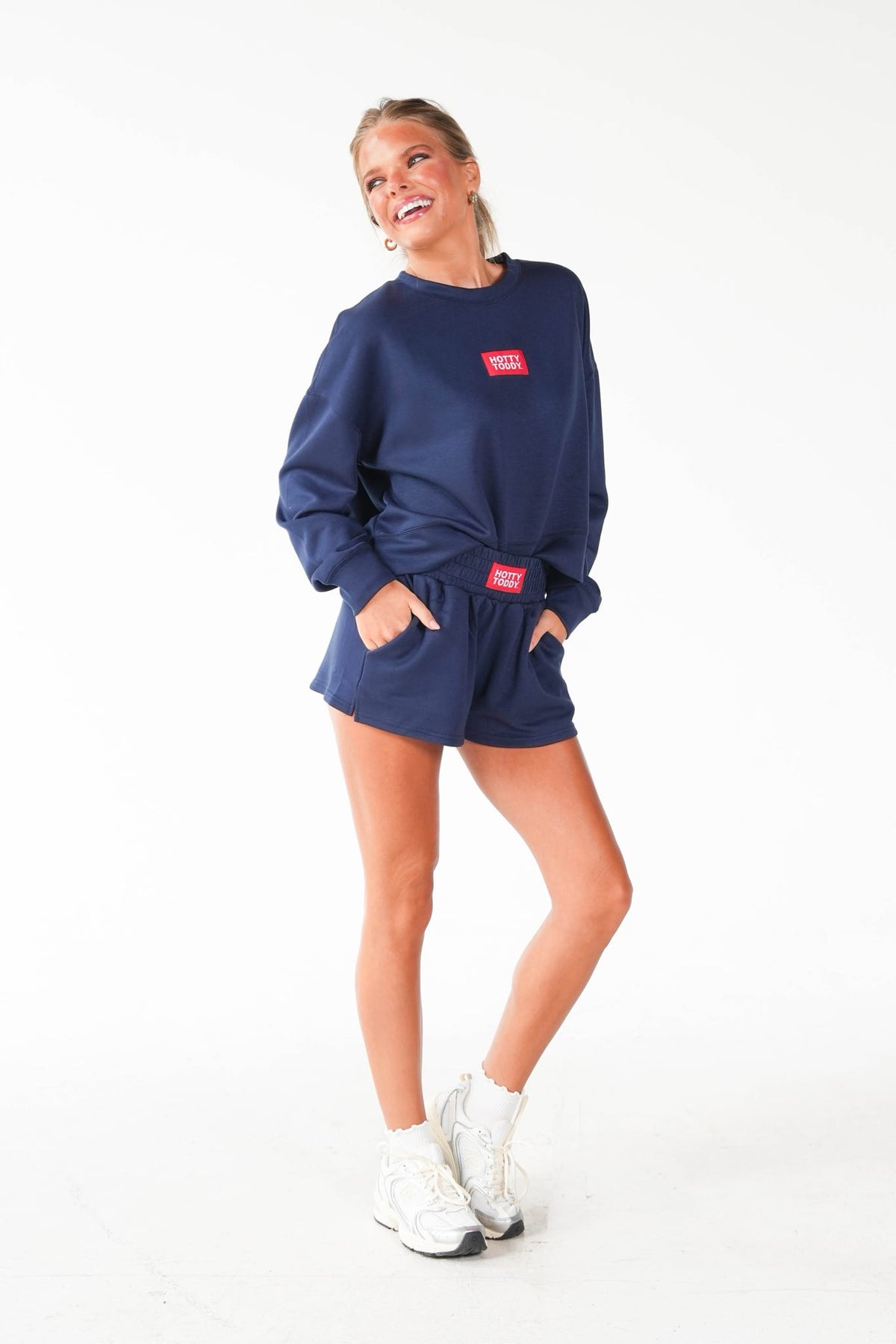A woman modeling a navy blue sweatshirt and matching shorts set with small red rectangular logo patches, paired with white sneakers, against a plain white studio background.