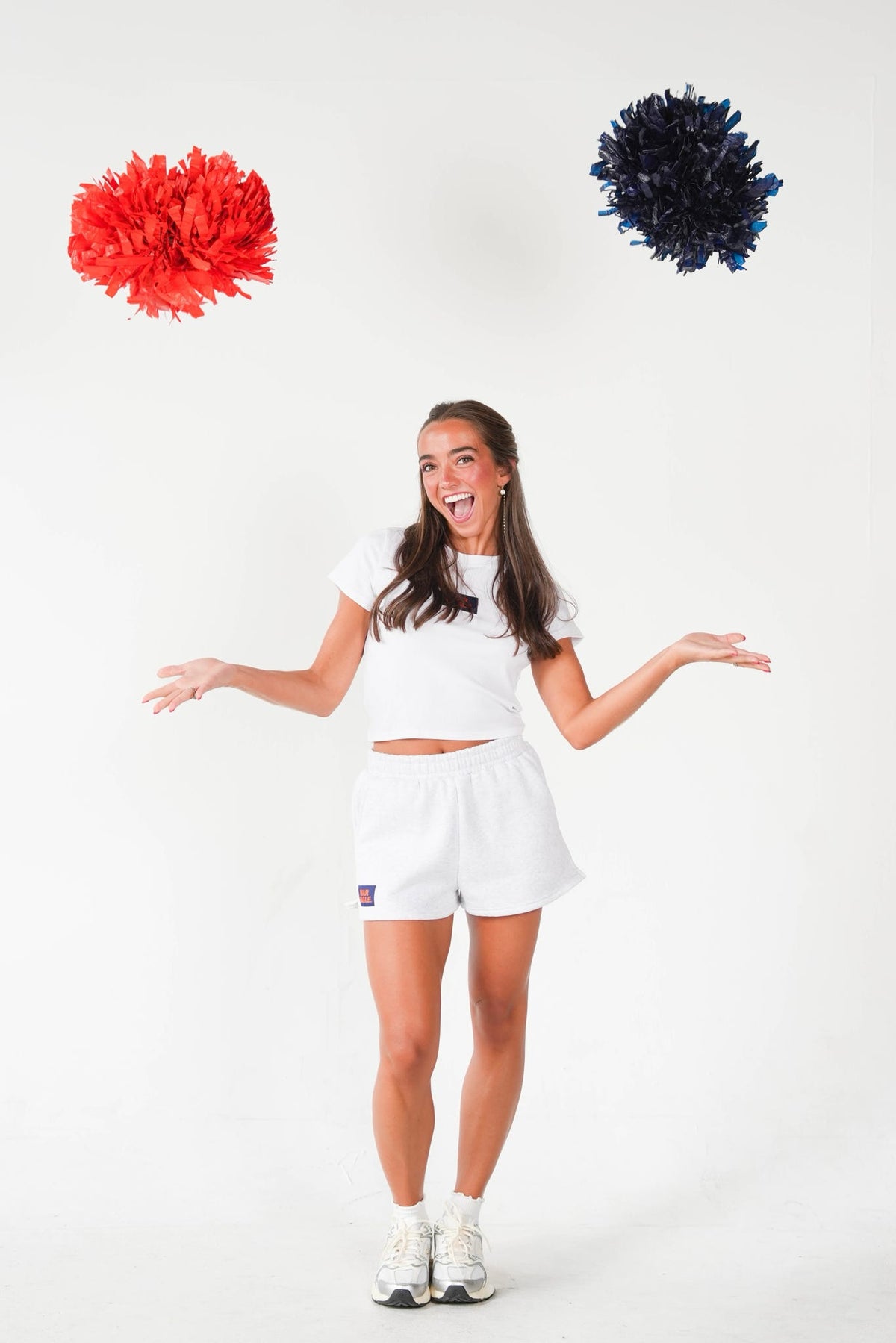 A woman in a white athletic crop top and shorts stands against a white studio background with her arms outstretched, looking up at two pom-poms floating in the air—one red and one dark blue/black.