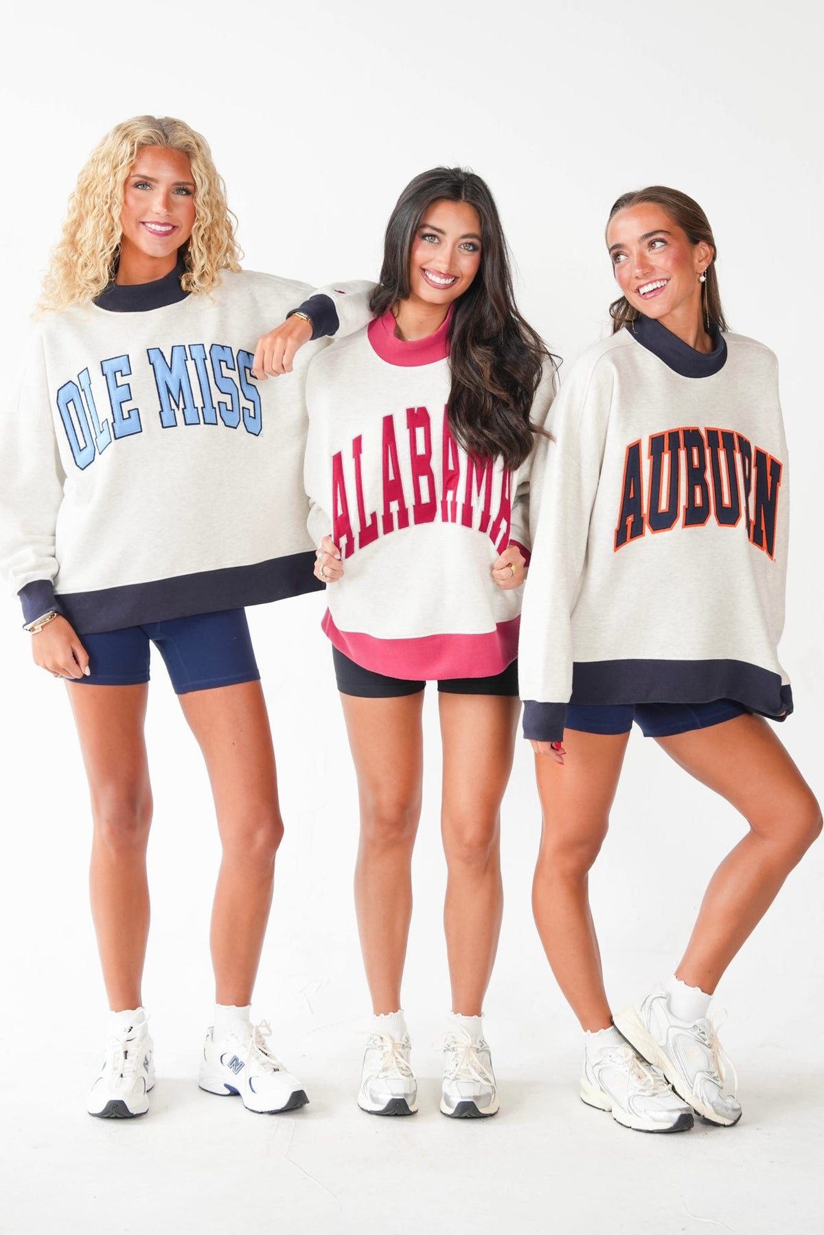 Three women modeling oversized collegiate sweatshirts with 'OLE MISS', 'ALABAMA', and 'AUBURN' lettering, paired with shorts and sneakers, against a plain white studio background.