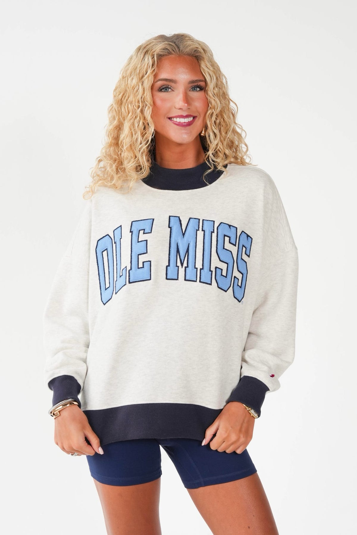 A woman modeling an oversized cream sweatshirt with 'OLE MISS' in light blue varsity letters outlined in navy across the chest, paired with navy shorts, against a plain white studio background.