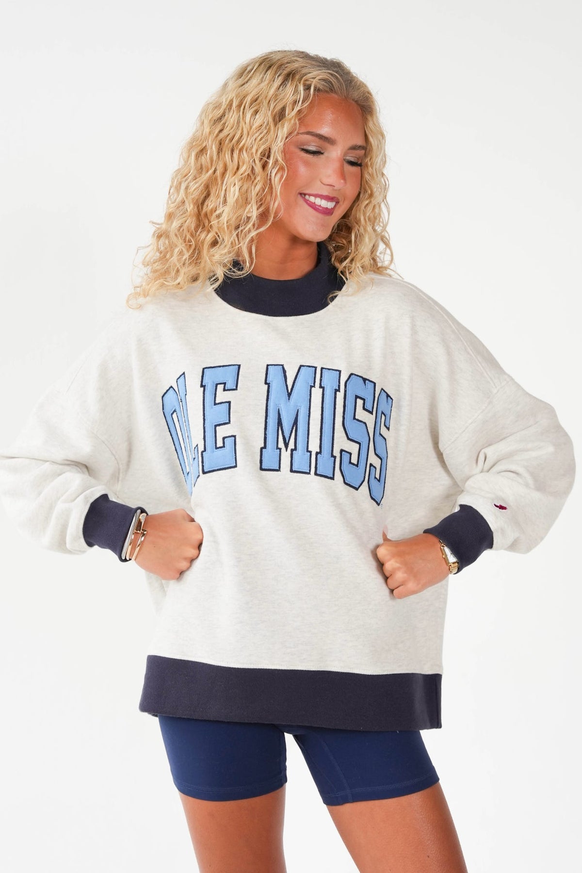A woman wearing an oversized cream sweatshirt with navy blue ribbed trim and 'OLE MISS' in light blue block letters across the chest, paired with navy blue bike shorts, against a plain white background.