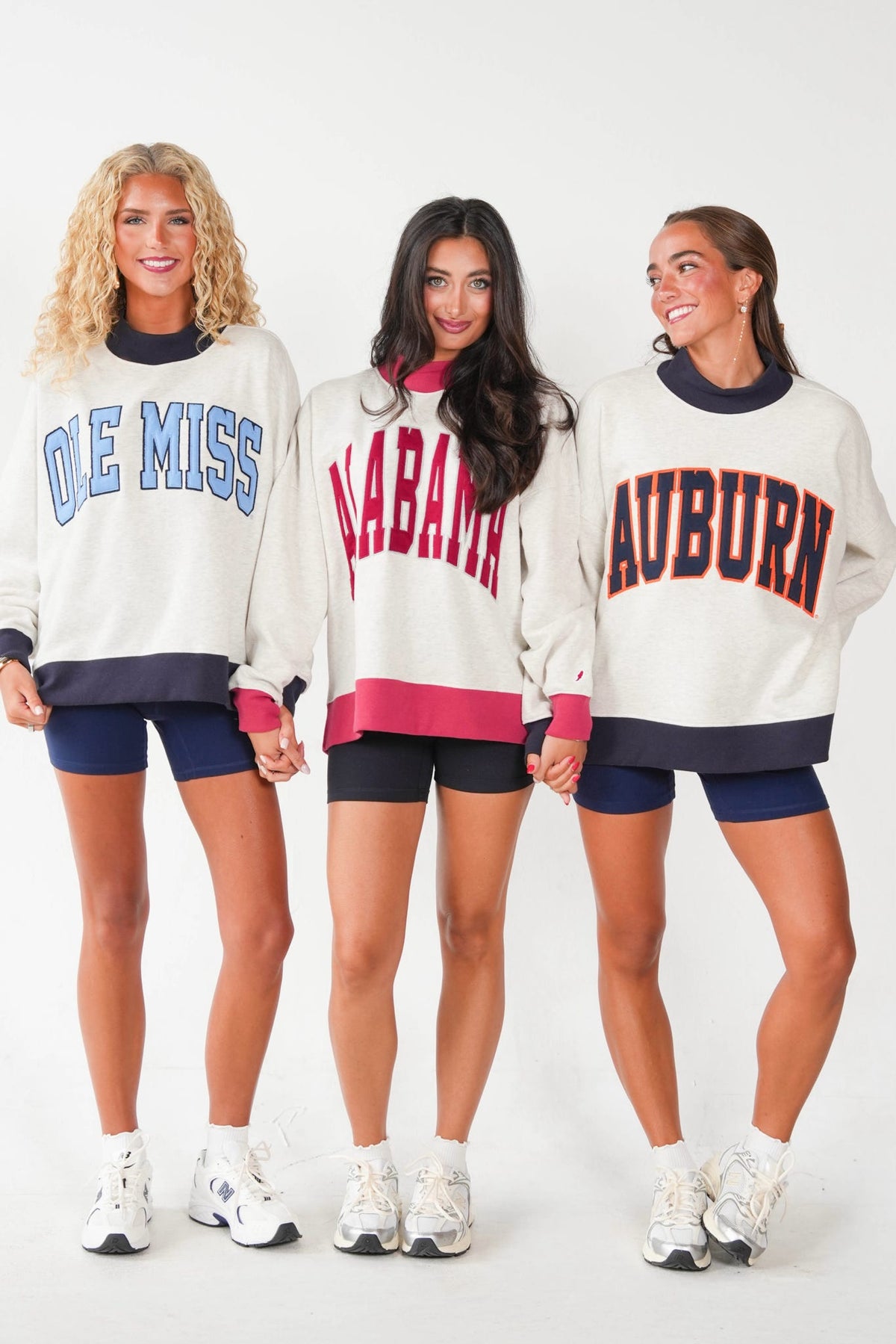 Three women standing side by side wearing matching cream-colored sweatshirts with collegiate lettering. The left sweatshirt says 'OLE MISS' in blue, the center says 'ALABAMA' in red, and the right says 'AUBURN' in red with navy trim.