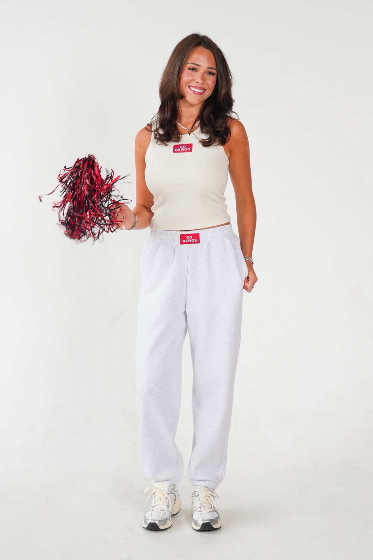 A woman modeling a white sleeveless crop top and matching white sweatpants, holding a red and black pom-pom against a plain white studio background.