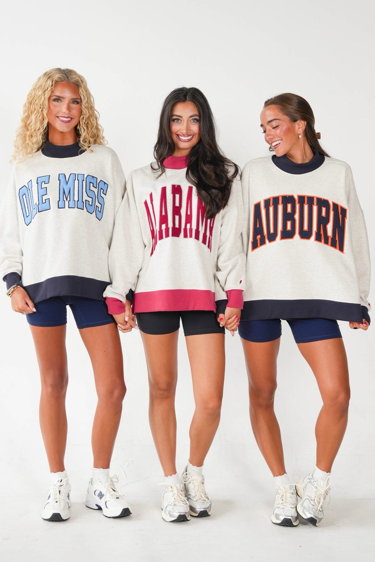 Three women modeling matching cream sweatshirts with collegiate lettering: 'Ole Miss' in blue, 'Alabama' in red, and 'Auburn' in red with navy trim. Each sweatshirt has a navy crew neck and hem, and the models are wearing navy shorts and white sneakers against a plain white studio background.