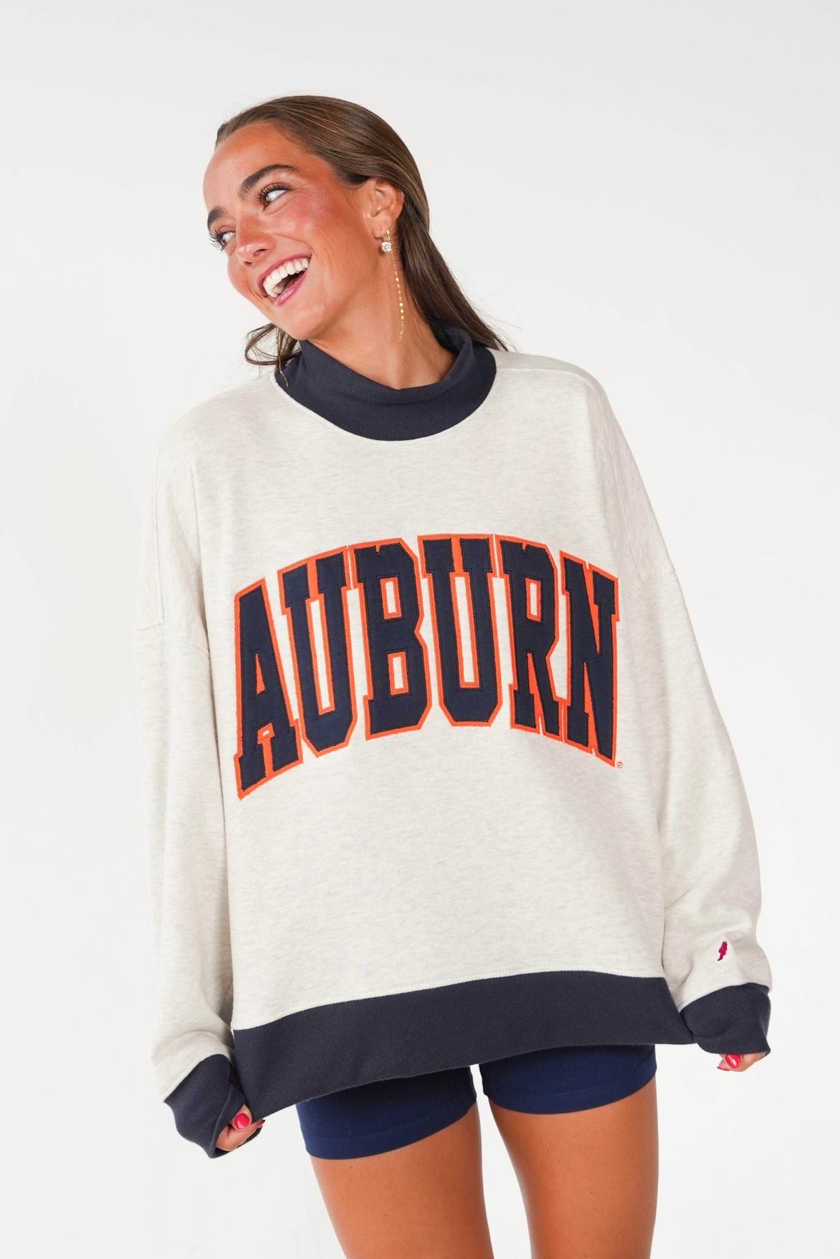 A woman wearing an oversized cream sweatshirt with 'AUBURN' printed across the front in navy and orange, paired with navy shorts, against a light gray studio background.