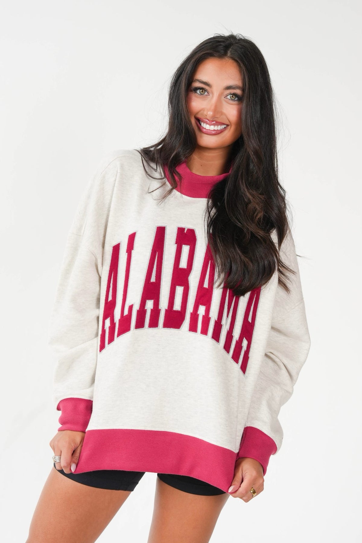 A woman modeling an oversized cream sweatshirt with bold magenta collegiate lettering that reads 'ALABAMA' across the front, paired with black shorts against a plain white studio background.