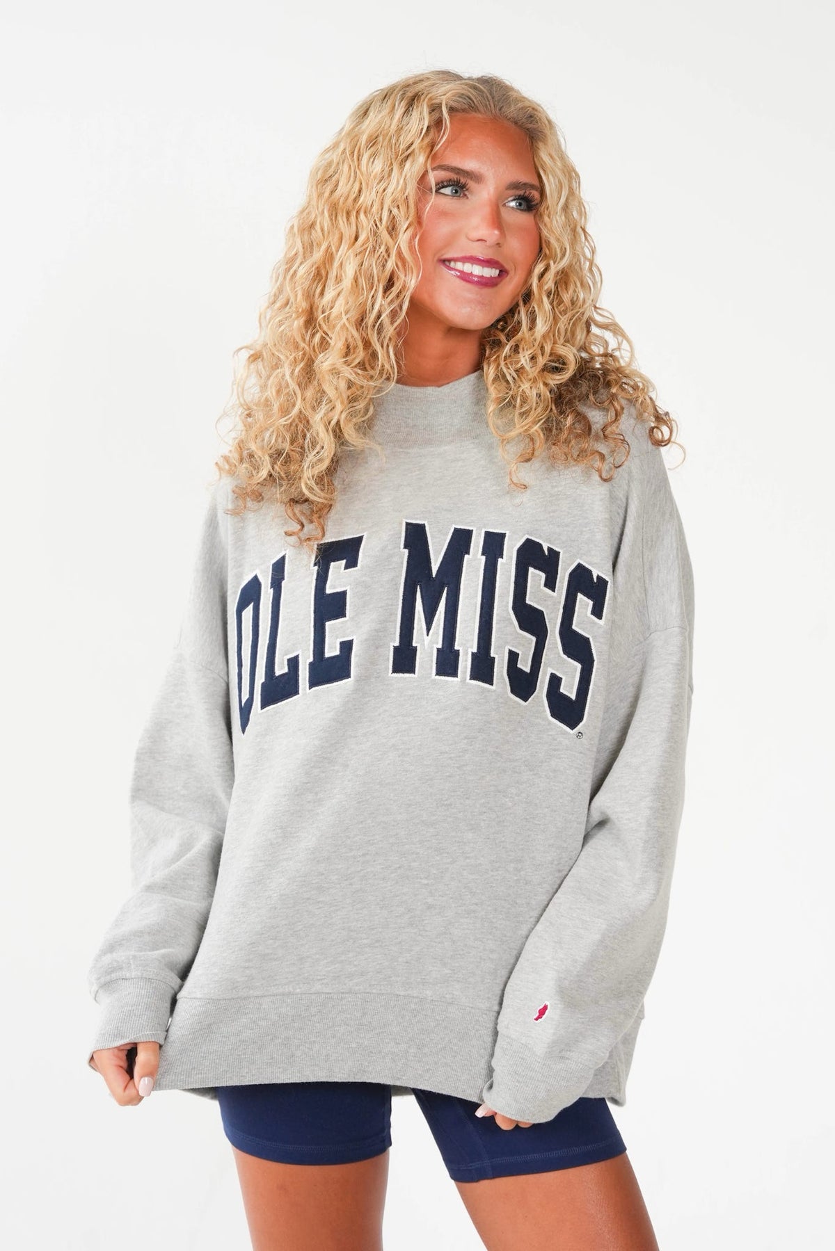 A woman wearing an oversized light gray sweatshirt with 'OLE MISS' printed across the chest in navy blue, paired with navy blue shorts, against a plain white background.