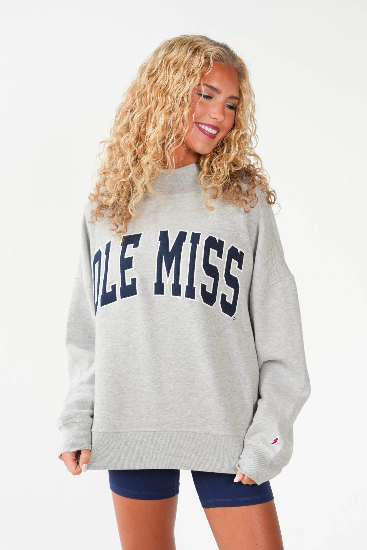 A woman with long curly blonde hair wearing an oversized light gray sweatshirt with 'OLE MISS' printed in navy blue across the chest, paired with navy blue bike shorts, against a plain white studio background.