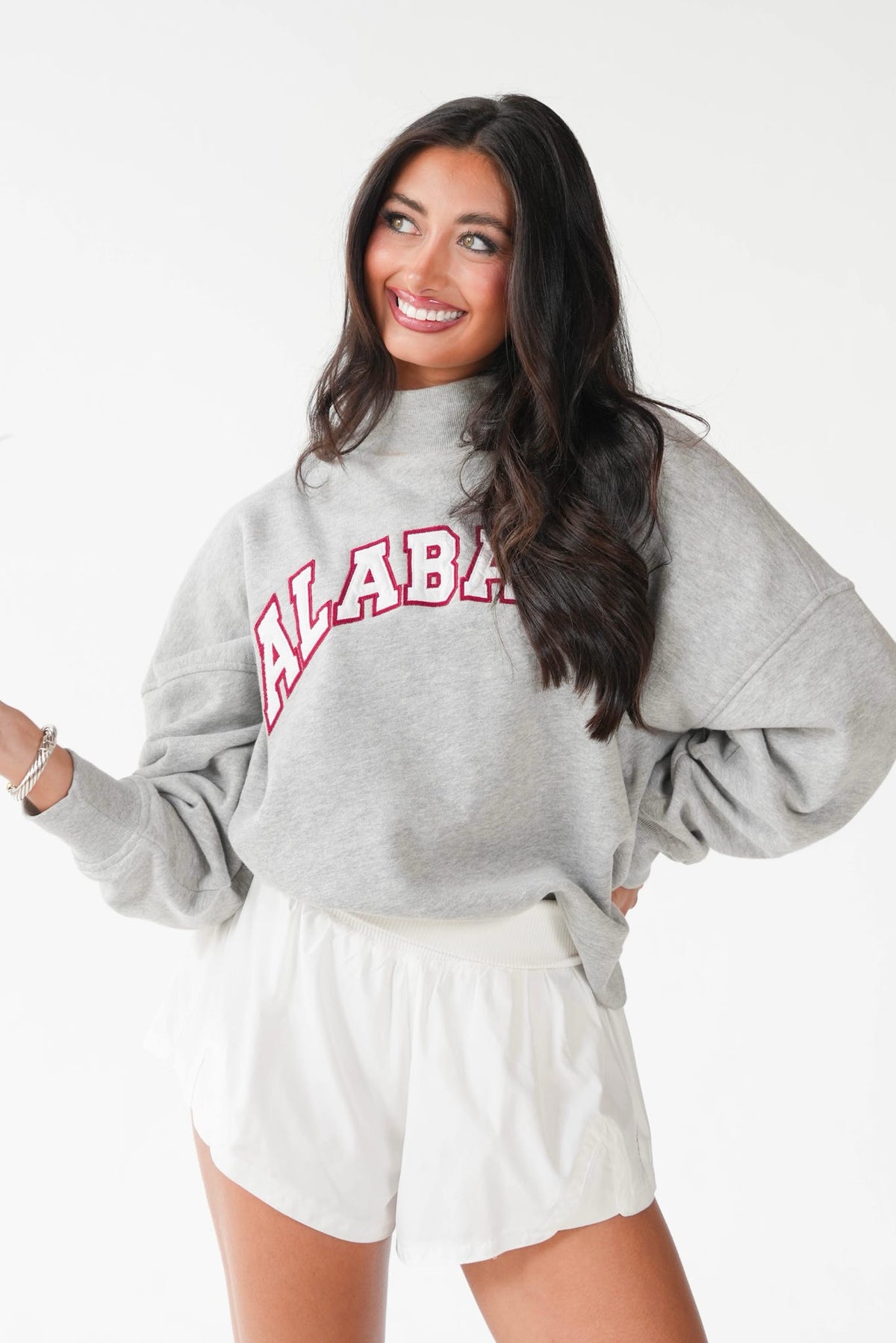 A woman modeling a light gray sweatshirt with 'ALABAMA' printed in red arched lettering across the chest, paired with white shorts, against a plain white background.