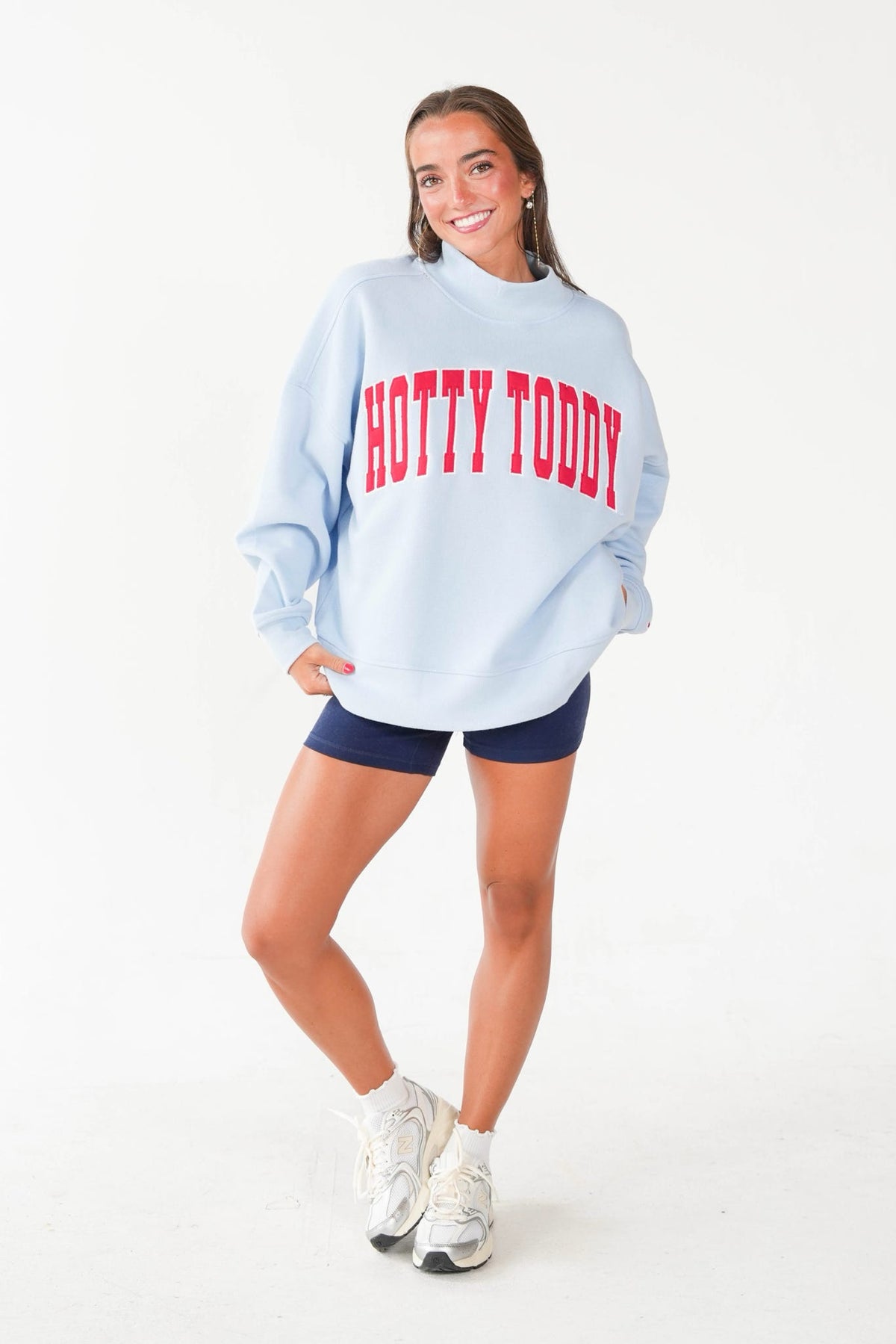 A woman modeling an oversized light blue sweatshirt with 'HOTTY TODDY' printed in red across the chest, paired with navy shorts and white sneakers against a plain white studio background.