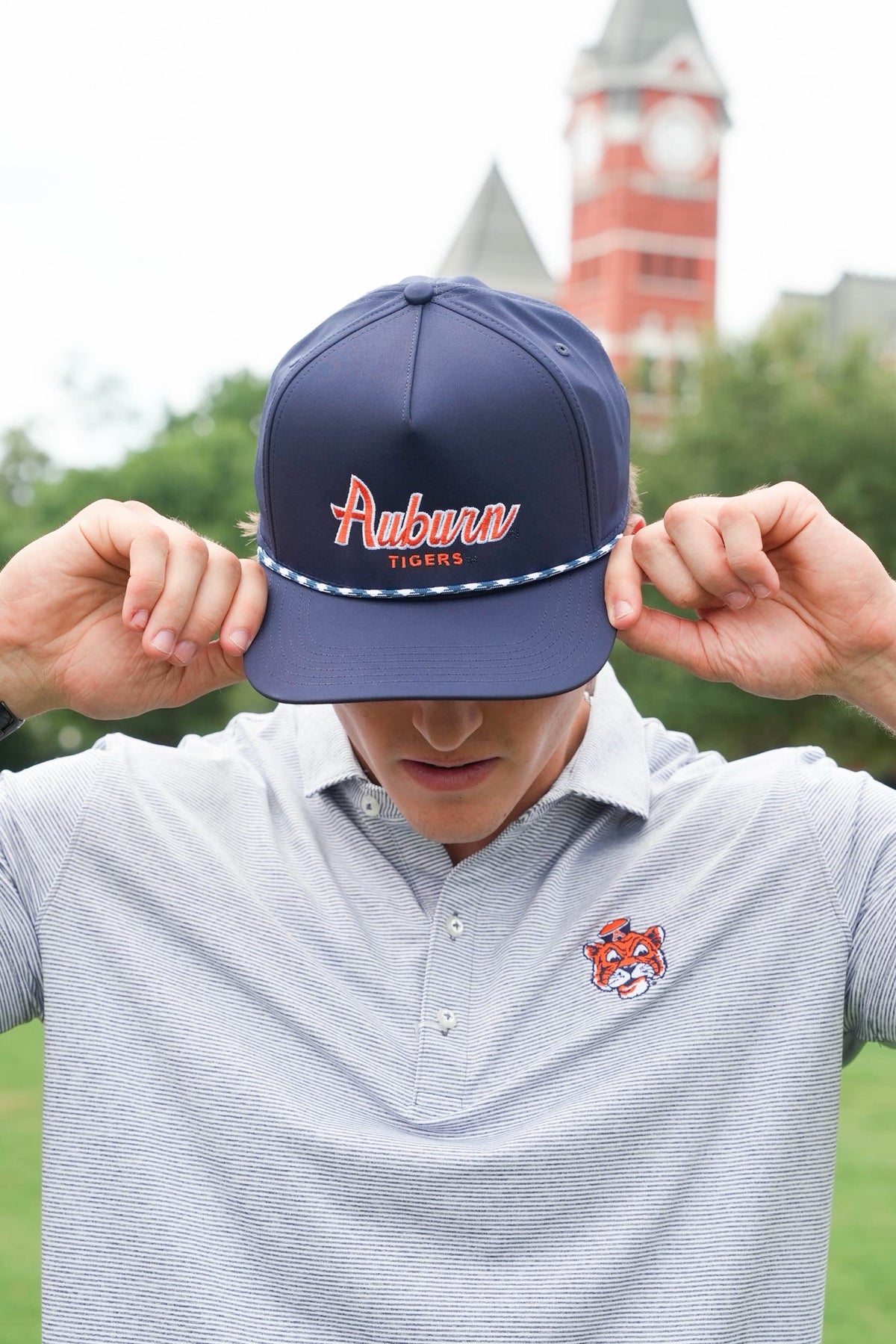 A man wearing a navy blue Auburn Tigers baseball cap with 'Auburn Tigers' embroidered on the front, holding the brim down over his eyes. He is wearing a gray polo shirt with an Auburn Tigers logo on the chest, with a blurred clock tower in the background.