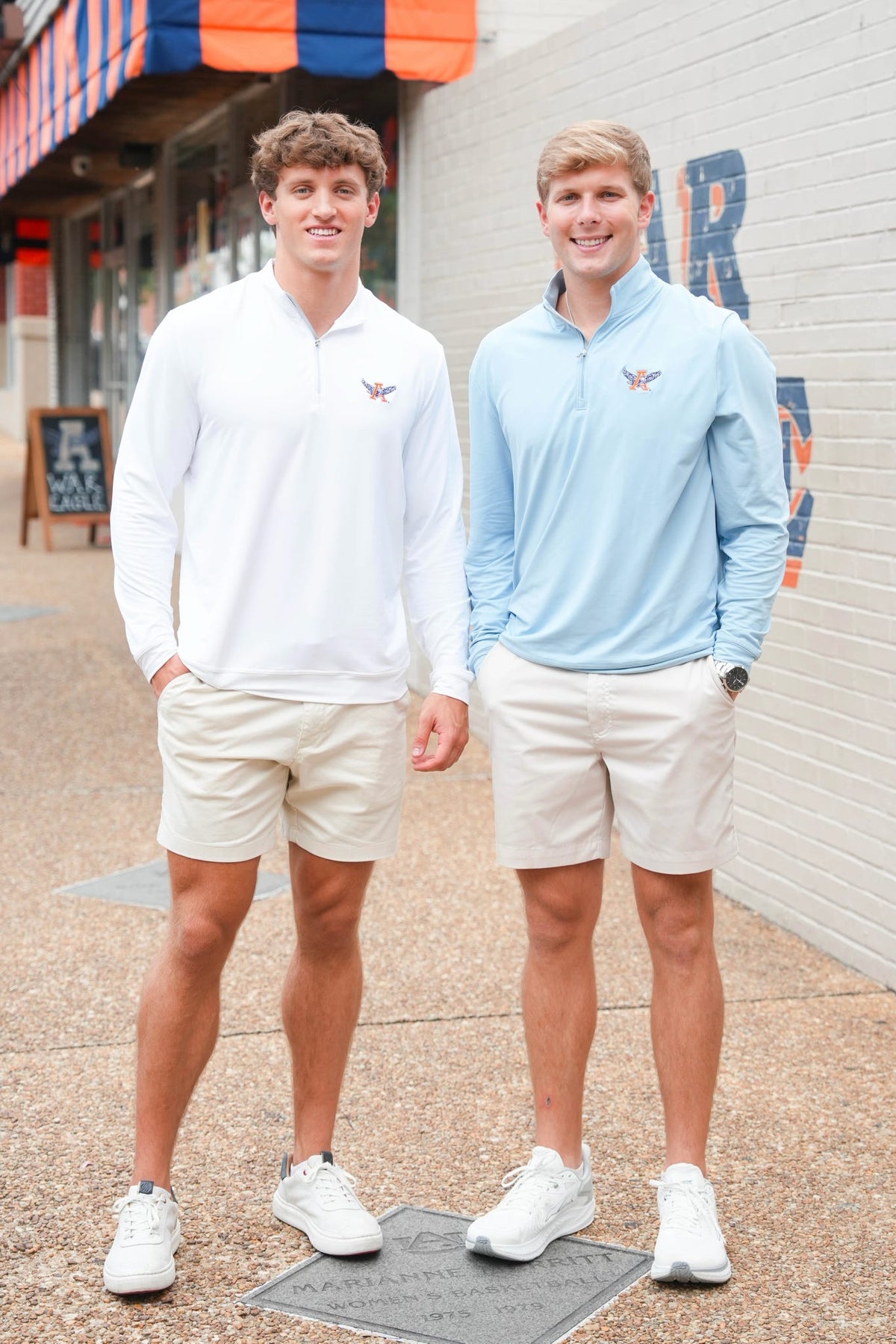 Two men standing outdoors on a sidewalk wearing matching long-sleeve quarter-zip performance shirts in white and light blue, paired with beige shorts and white sneakers.