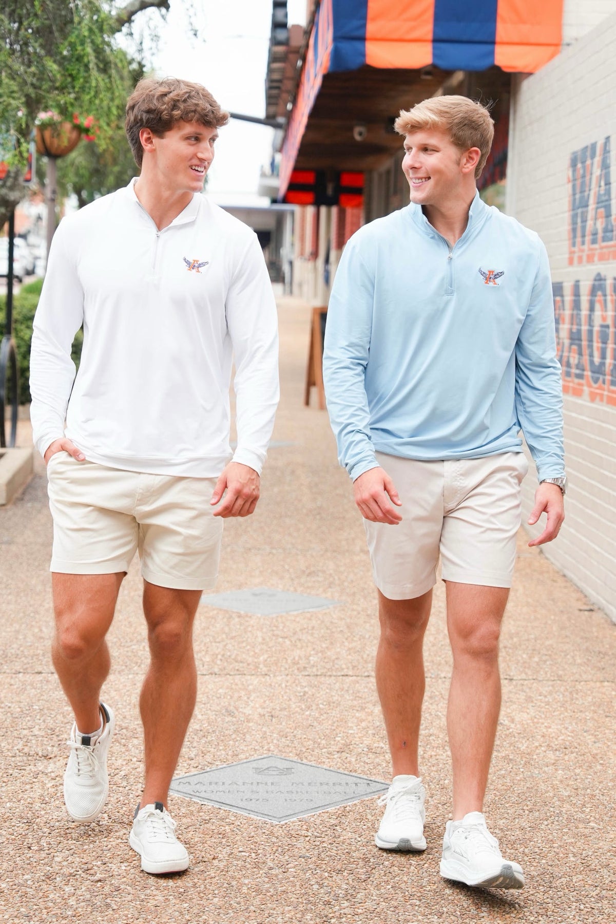 Two men walking outdoors on a paved street wearing matching polo shirts and shorts. The man on the left wears a white long-sleeve polo with a small chest logo and beige shorts, while the man on the right wears a light blue long-sleeve polo with a small chest logo and beige shorts.