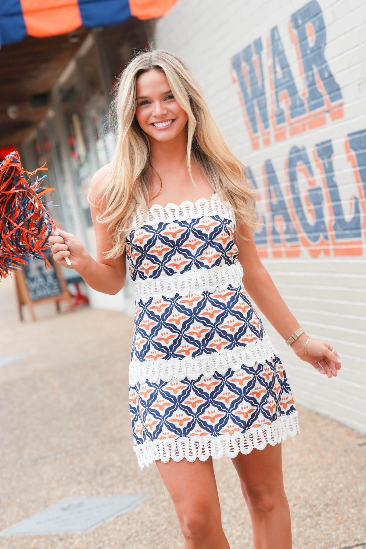 A woman wearing a blue, white, and orange patterned crochet strapless mini dress with white lace trim, standing outdoors in front of a white brick wall with blue and orange lettering.