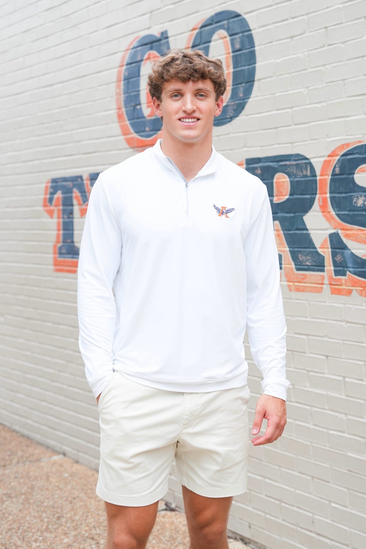 A young man wearing a white long-sleeve quarter-zip pullover and cream shorts, standing in front of a white brick wall with large blue and orange lettering.