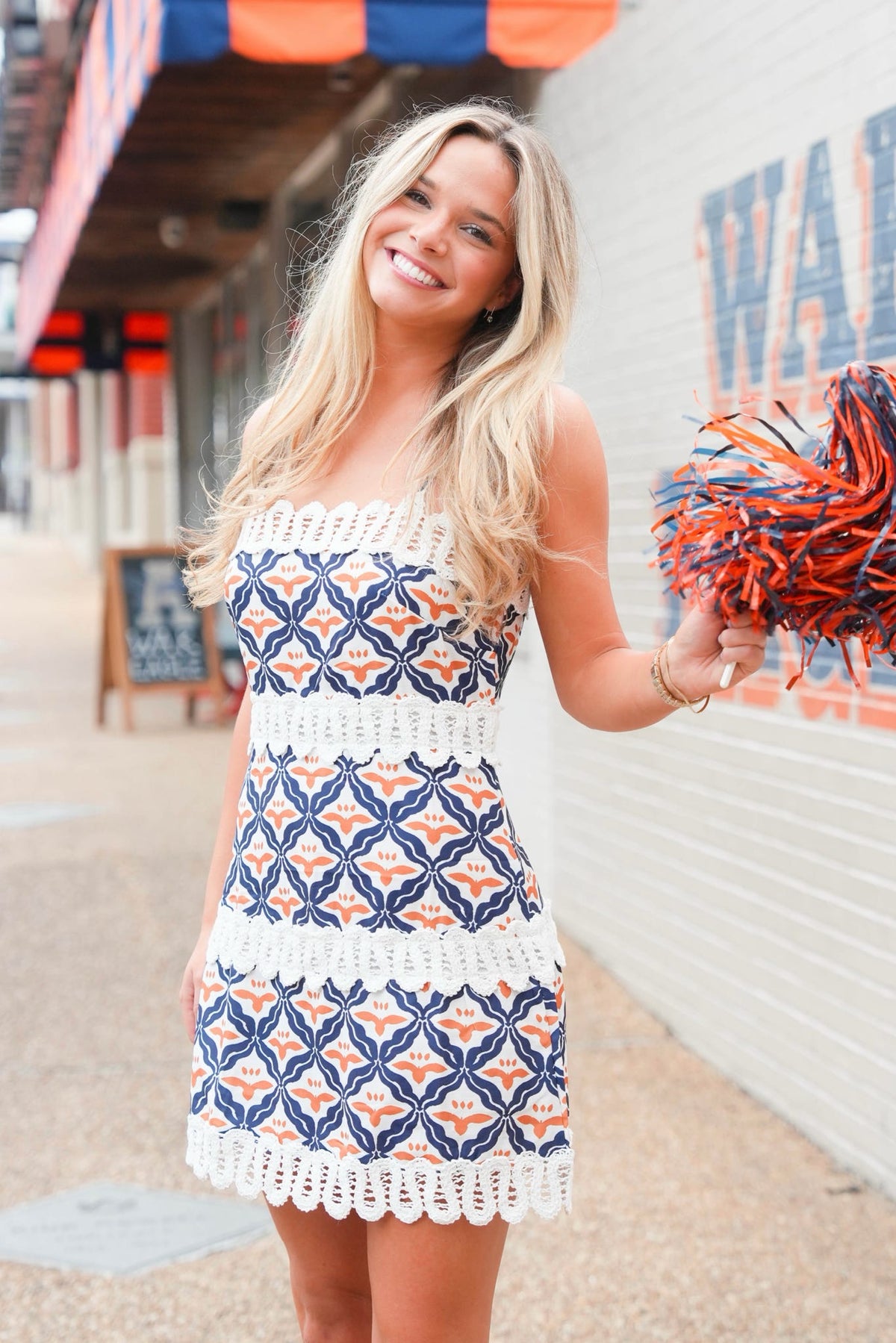A smiling blonde woman wearing a sleeveless crochet mini dress with a navy blue, white, and orange geometric pattern, holding a matching orange and navy cheer pom-pom in an outdoor setting.