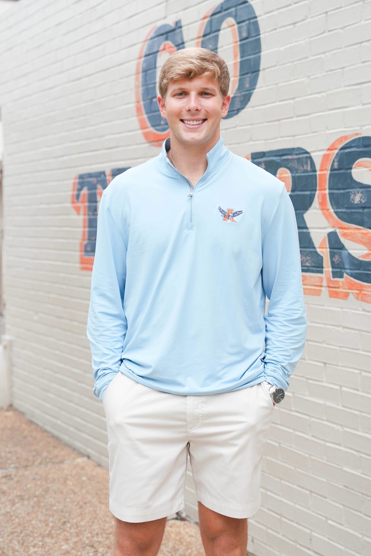A smiling young man stands outdoors against a white brick wall with large painted letters, wearing a light blue long-sleeve quarter-zip pullover with a small bird logo on the left chest and off-white shorts.