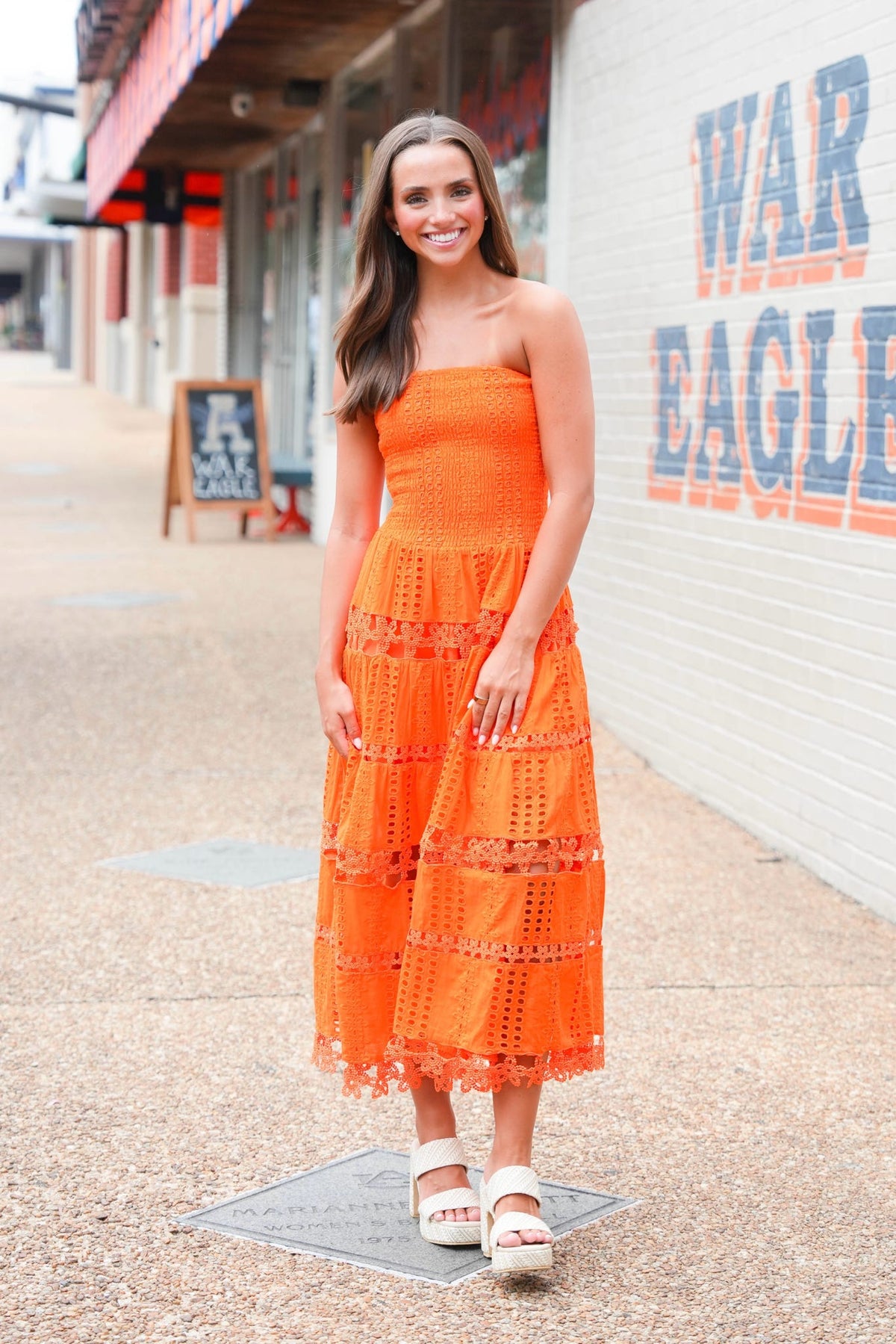 A woman wearing a bright orange strapless crochet midi dress and white platform sandals stands on a sidewalk in front of a white brick wall with 'WAR EAGLE' painted on it.