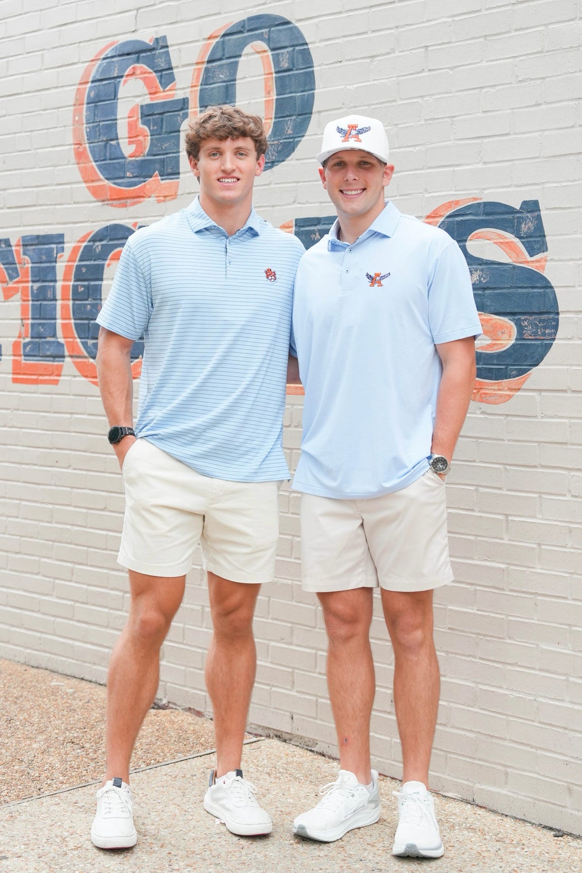 Two men standing outdoors in front of a brick wall with large painted letters, wearing matching light blue polo shirts, beige shorts, and white sneakers. One man wears a white cap with a logo.