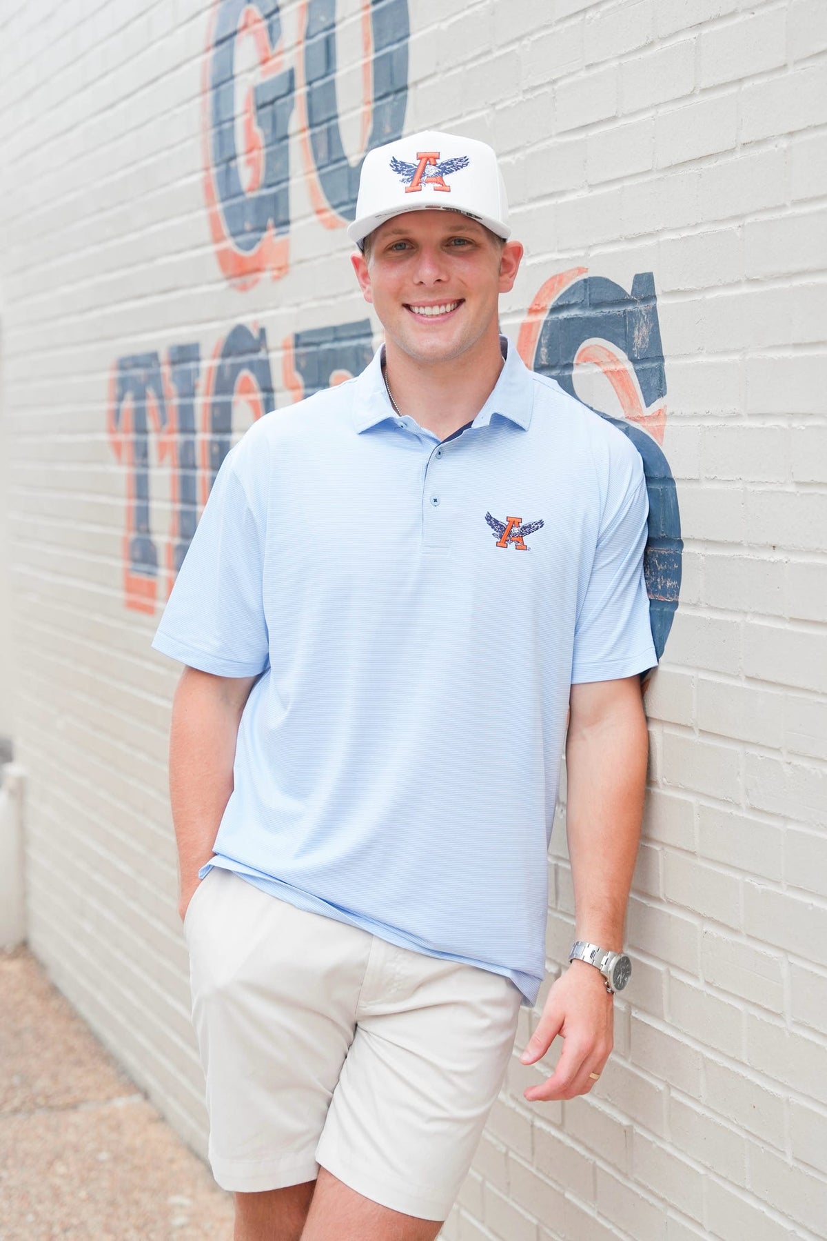 A man wearing a light blue polo shirt with a small embroidered logo on the left chest, a matching white cap, and beige shorts, standing against a white brick wall with painted lettering.