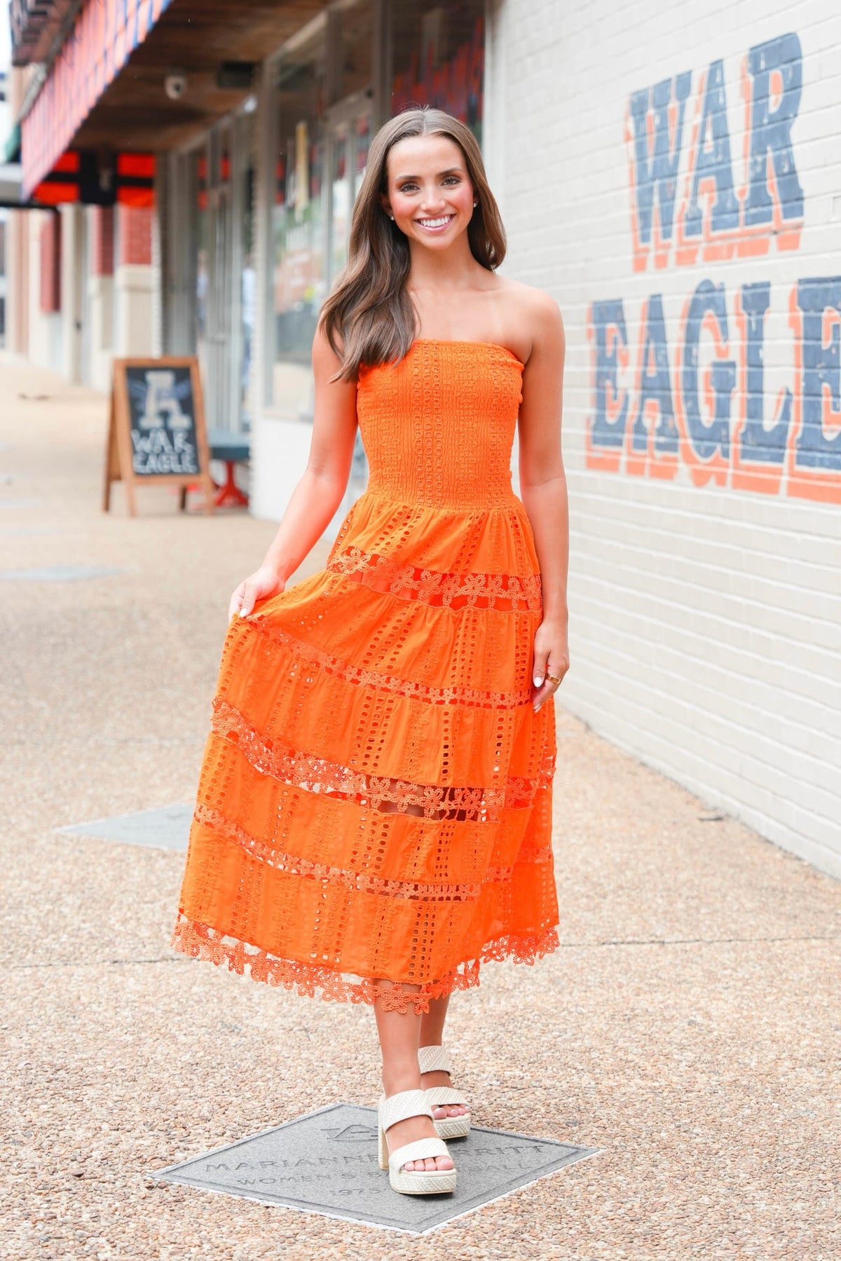 A woman wearing a bright orange strapless eyelet midi dress and white platform sandals stands on a sidewalk in front of a white brick wall with 'WAR EAGLE' painted on it.