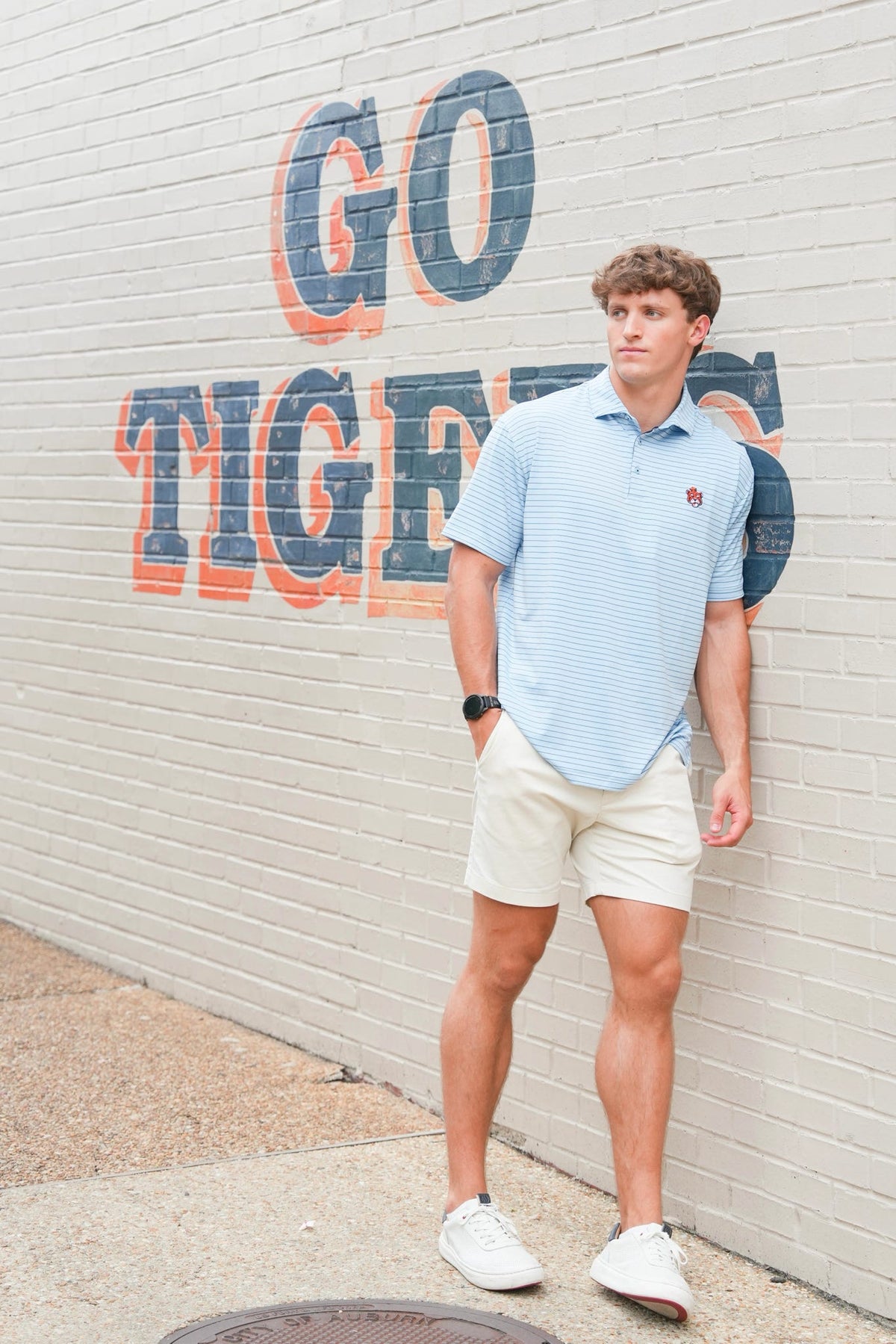 A man wearing a light blue and white striped polo shirt, beige shorts, and white sneakers stands against a white brick wall with a large 'GO TIGERS' mural.