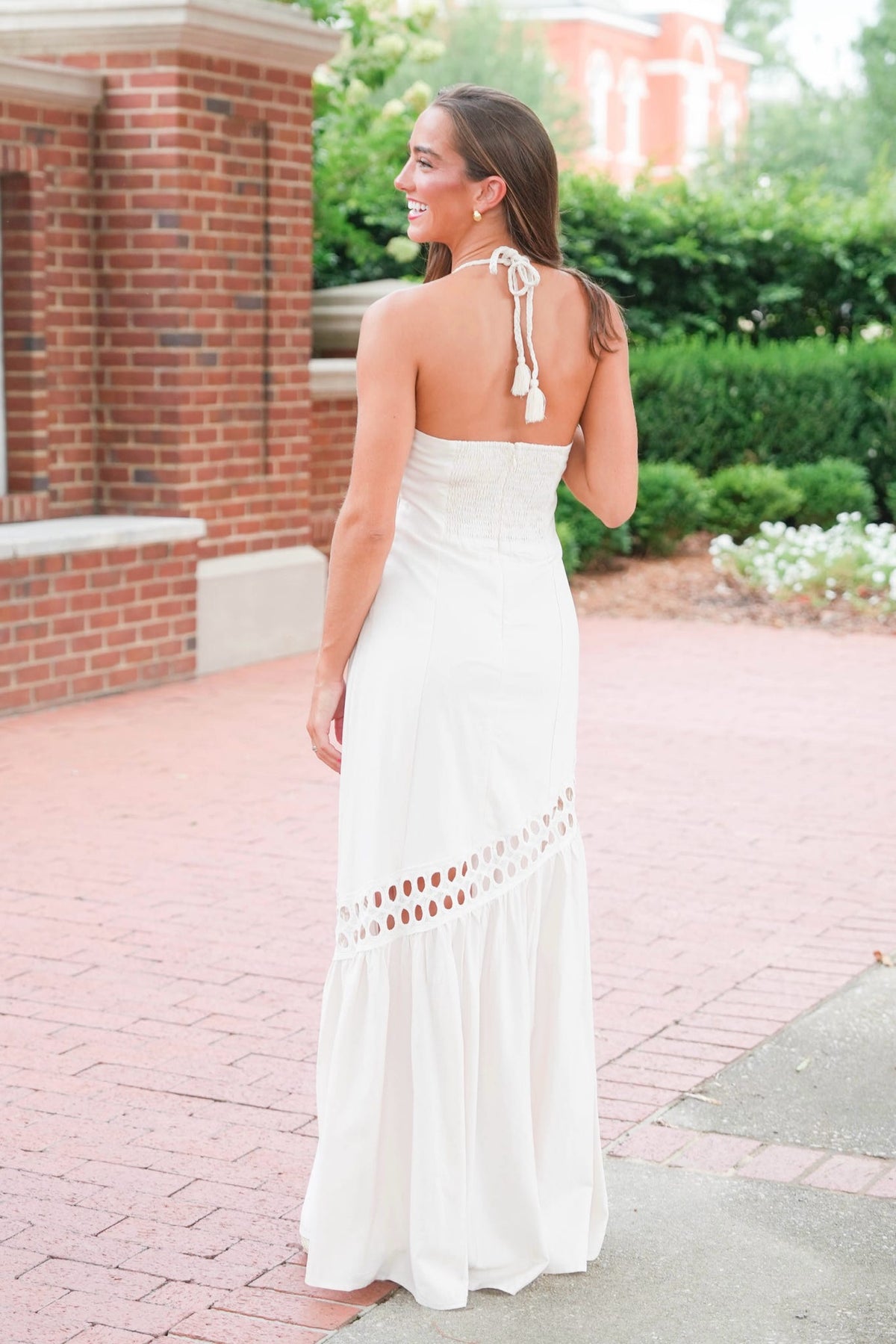 A woman wearing a strapless white maxi dress with tie-back halter details and a ruffled hem, photographed outdoors on a brick walkway with greenery and a brick building in the background.