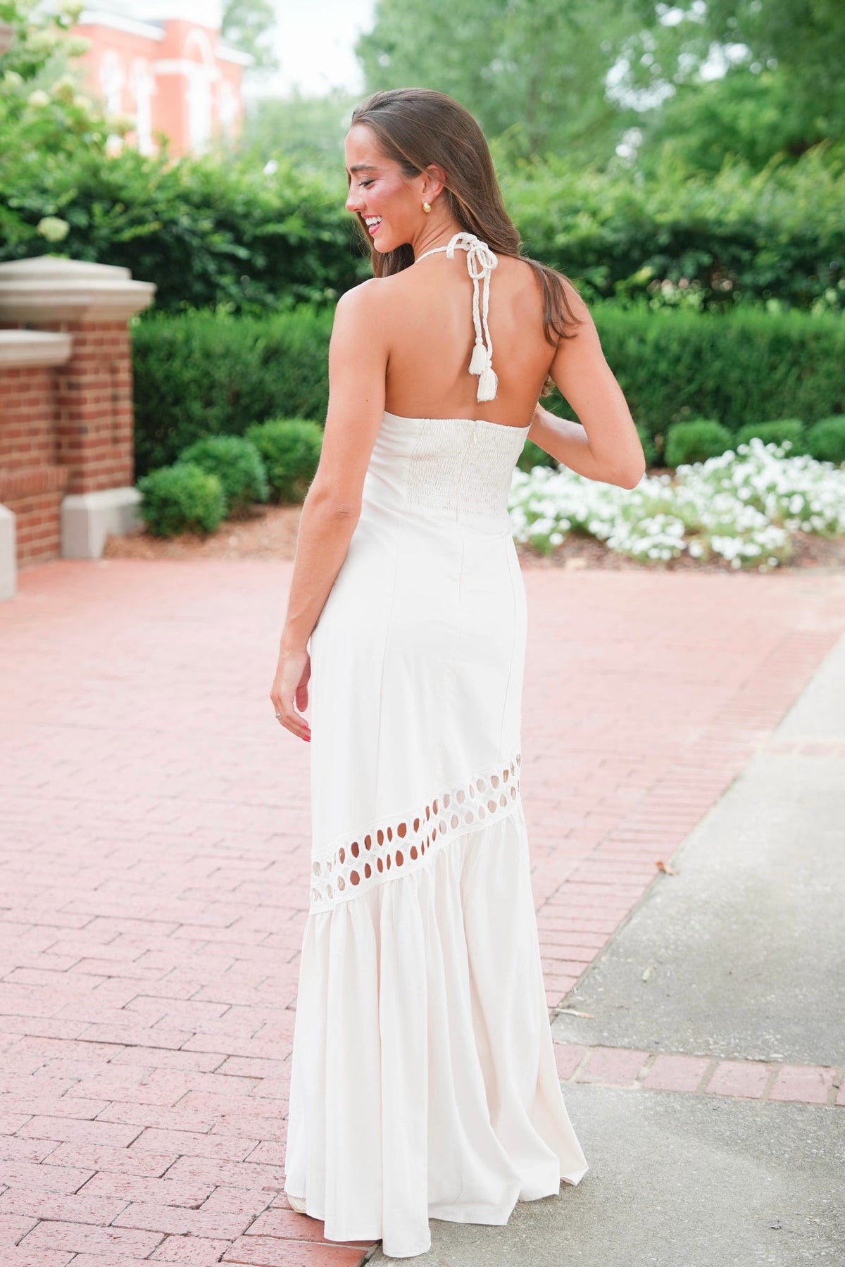 A woman wearing a sleeveless white maxi dress with tie-back straps and a ruffled hem, photographed outdoors on a brick walkway with greenery in the background.
