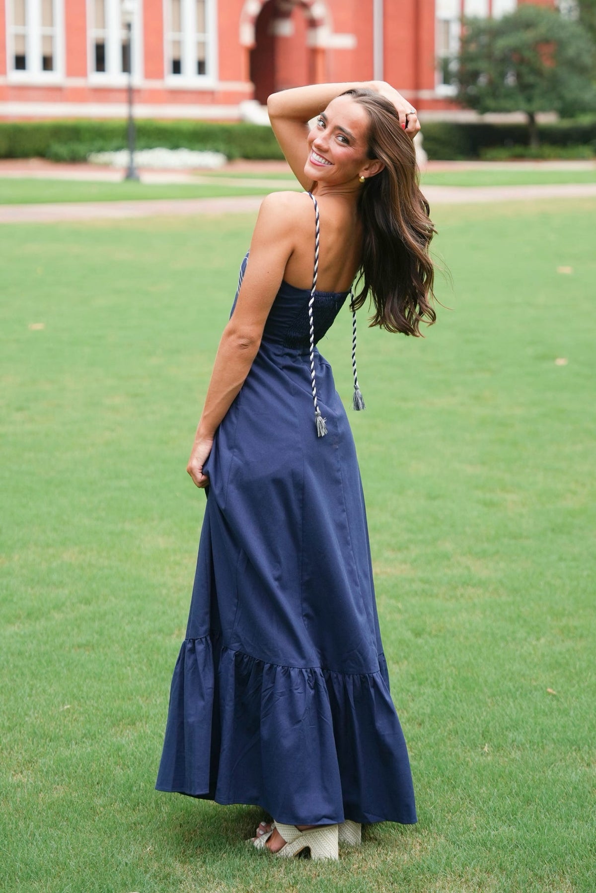 A woman wearing a navy blue strapless maxi dress with a braided back detail, standing on a green lawn with a red brick building in the background.