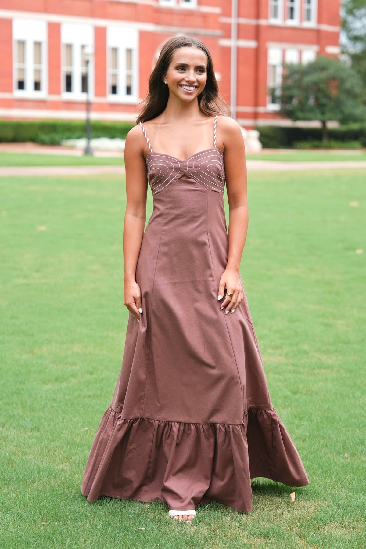 A woman wearing a floor-length mauve-brown spaghetti strap dress stands on a green lawn in front of a red brick building.