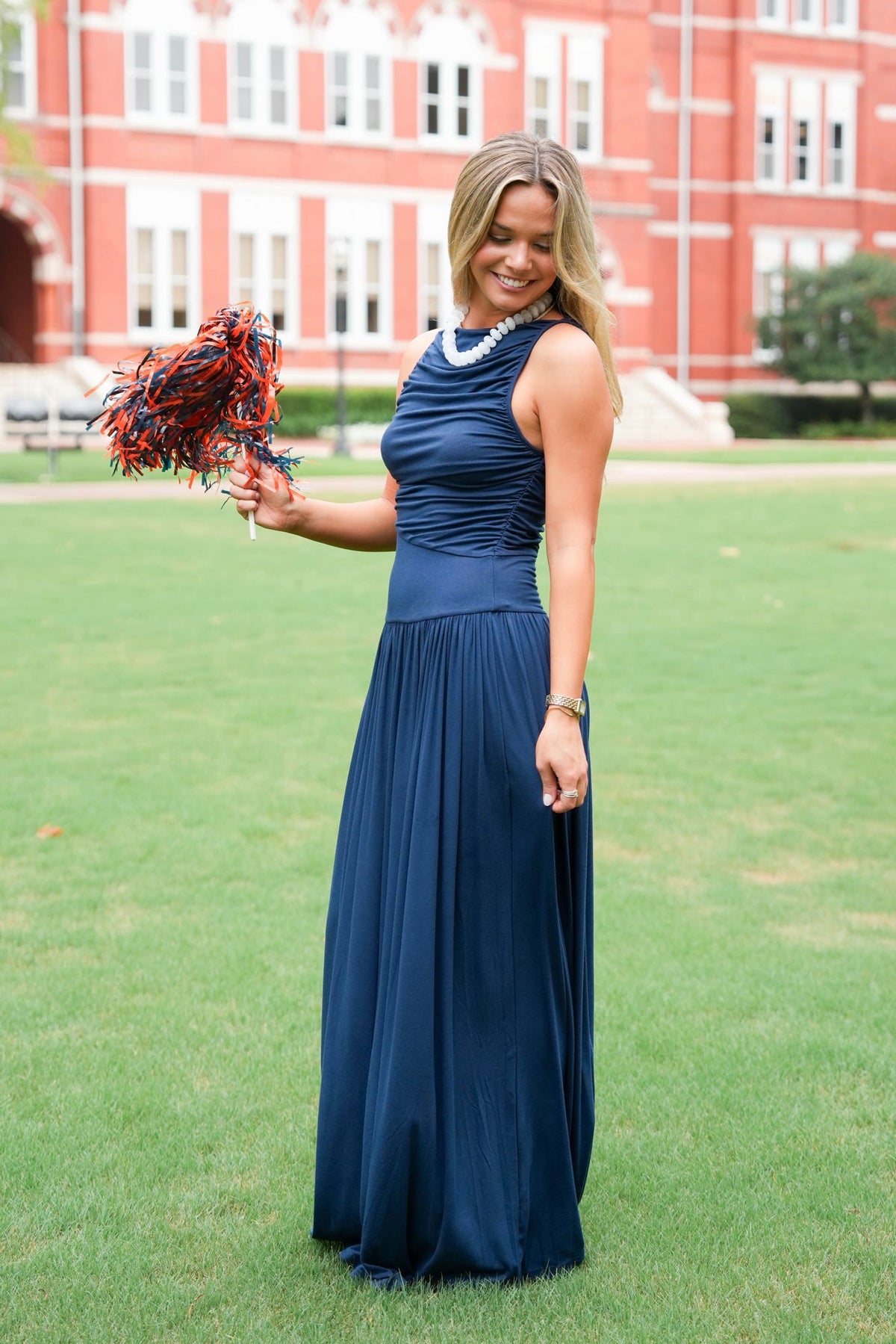 A woman stands on a grassy lawn in front of a large red brick building, wearing a sleeveless navy blue maxi dress with a pleated bodice and flowing skirt. She is holding a red and blue pom-pom.