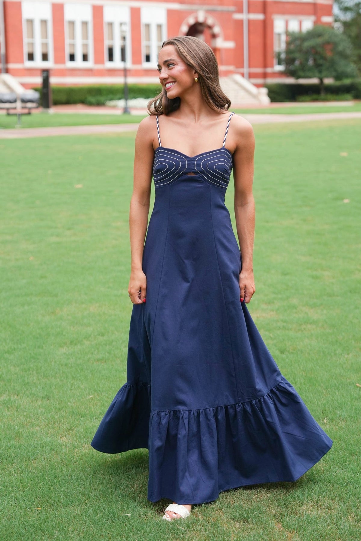 A woman wearing a navy blue maxi dress with a sweetheart neckline and embroidered detailing on the bodice, standing on a green lawn with a brick building in the background.