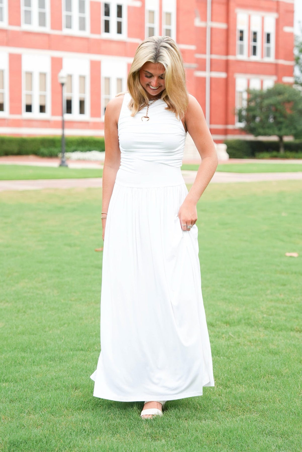 A woman wearing a sleeveless white maxi dress stands on a green lawn in front of a large red brick building.