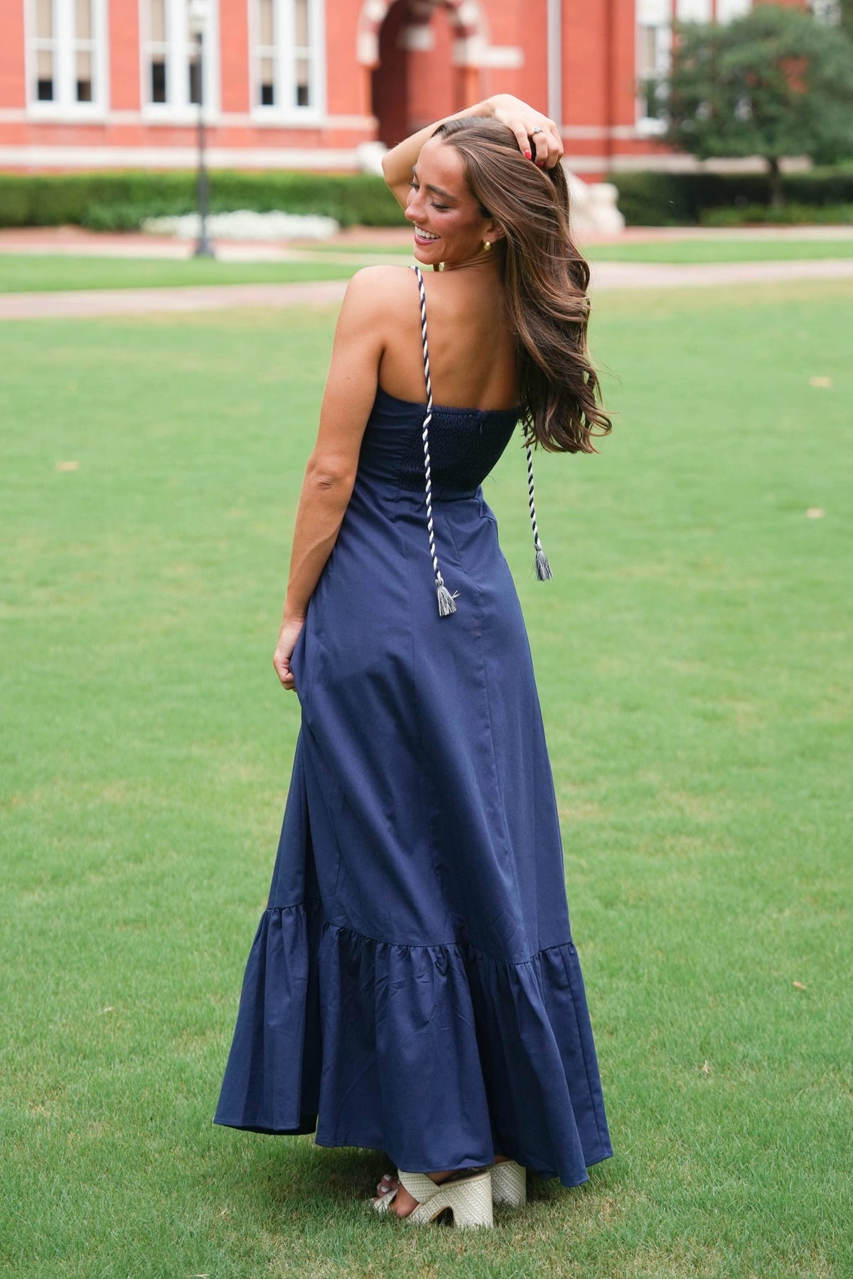 A woman stands on a green lawn wearing a navy blue strapless maxi dress with a tie-back design, photographed outdoors with a brick building in the background.