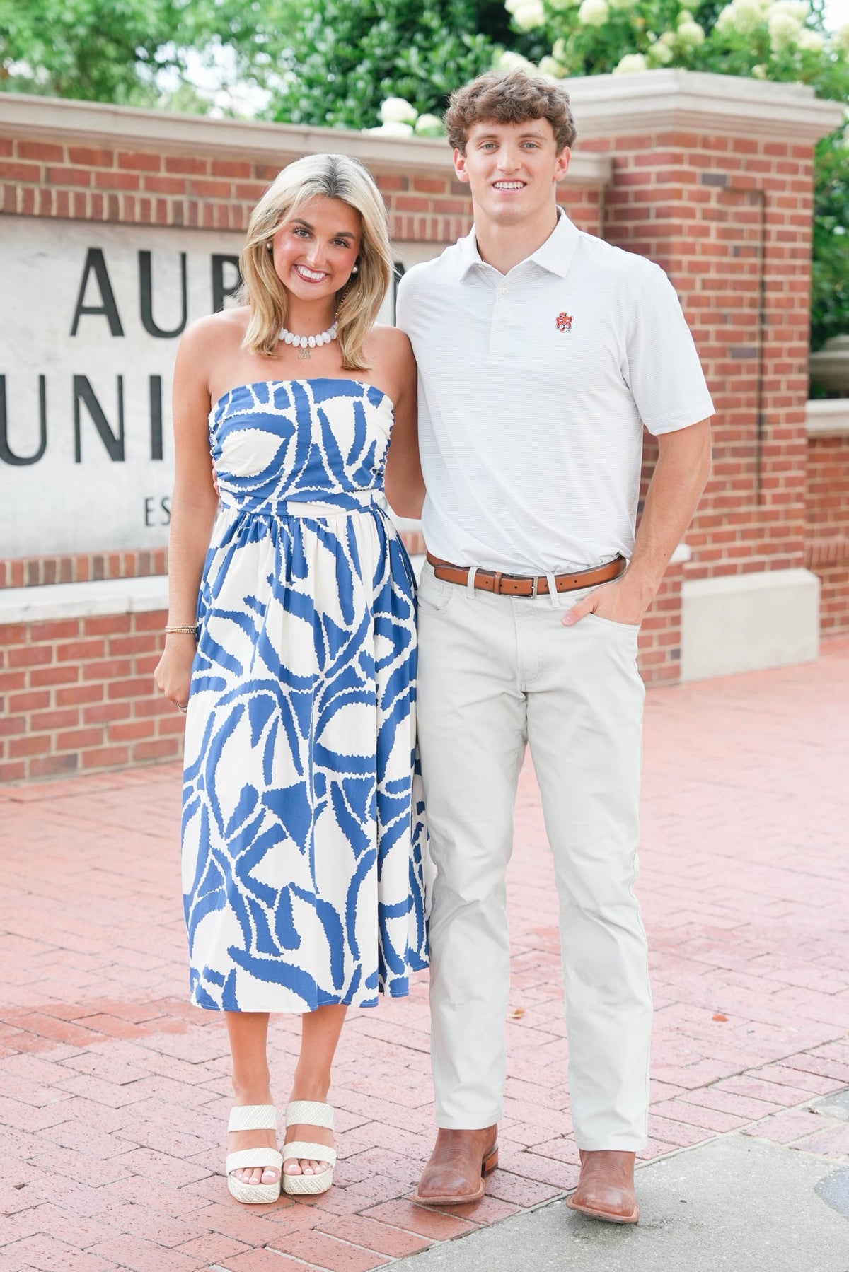 A couple standing outdoors in front of a brick wall and sign. The woman wears a strapless blue and white patterned midi dress, and the man wears a white polo shirt, cream pants, and brown shoes.