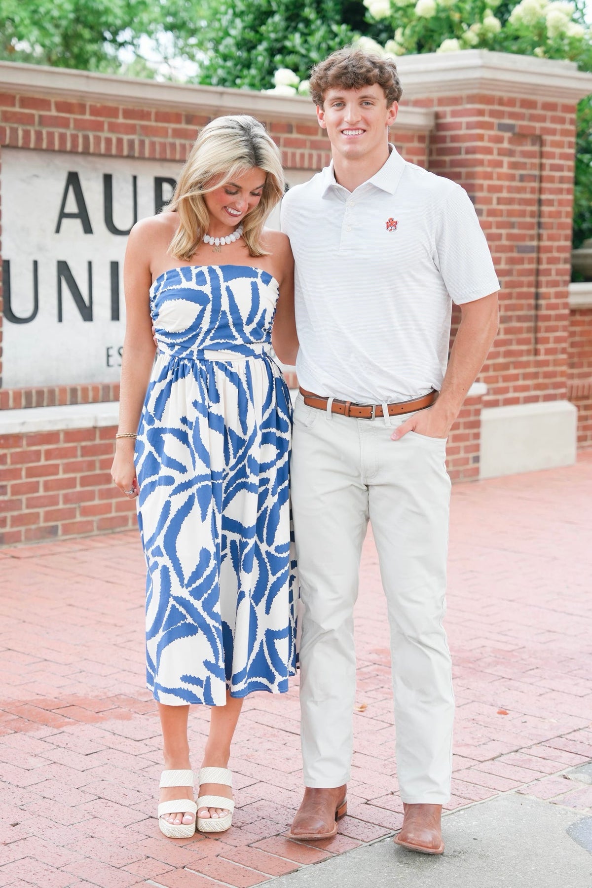 A couple standing outdoors in front of a brick wall with a sign. The woman wears a strapless white dress with a bold blue abstract leaf pattern, and the man wears a white polo shirt, cream pants, and brown shoes.