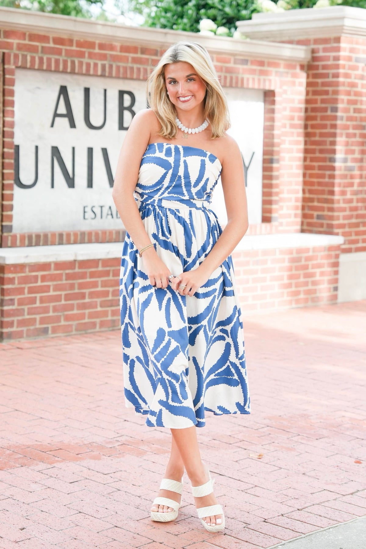 A woman modeling a strapless blue and white patterned midi dress with a matching belt, standing outdoors in front of a brick wall with a 'Auburn University' sign.