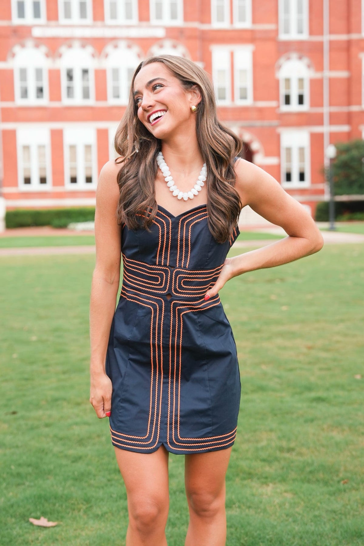 A woman wearing a navy blue sleeveless dress with orange geometric stripe patterns and a white pearl necklace, standing on a green lawn in front of a red brick building.
