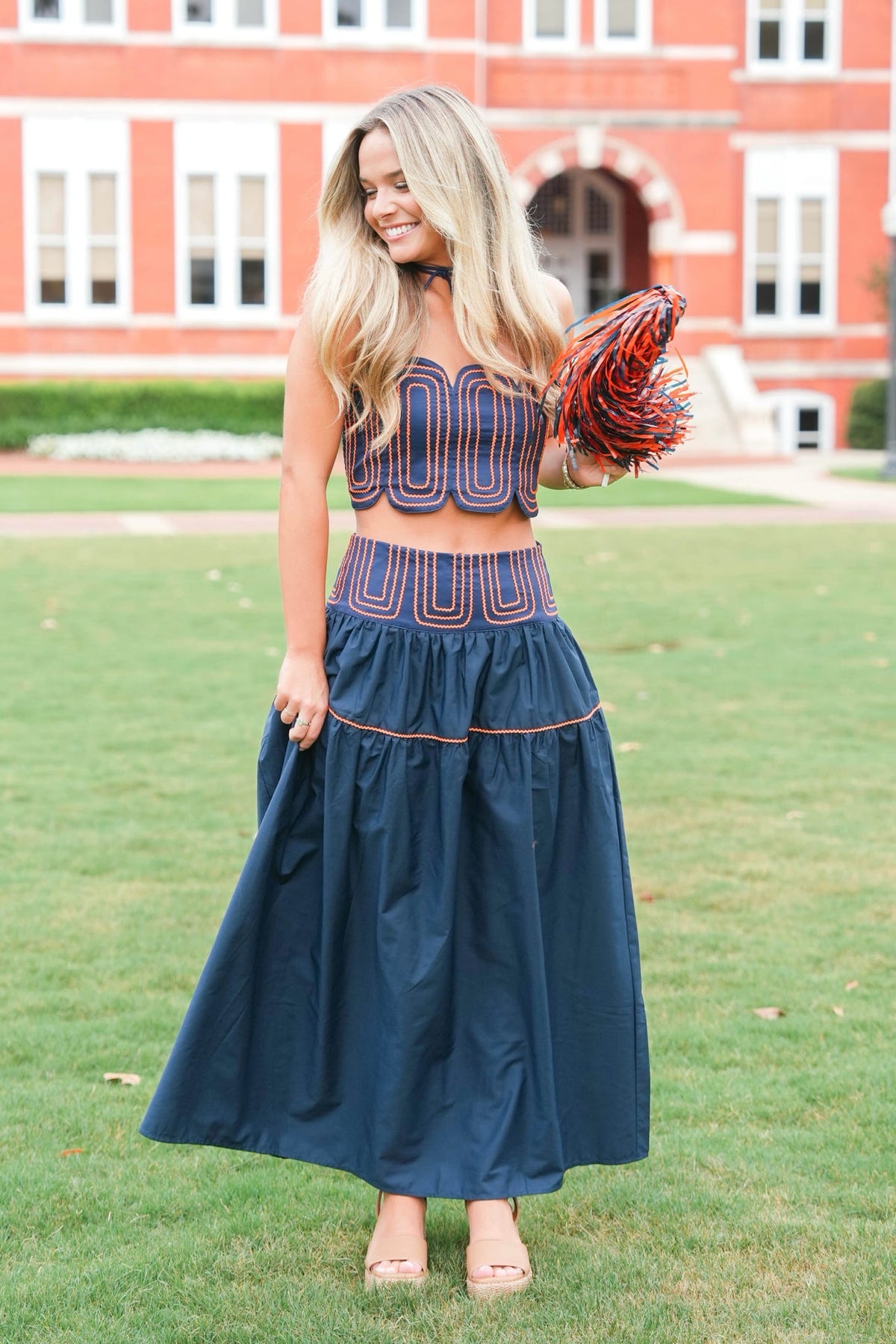 A woman stands on a grassy lawn in front of a red brick building, wearing a navy blue two-piece set with geometric patterned crop top and matching flared skirt, holding a red and navy pom-pom.