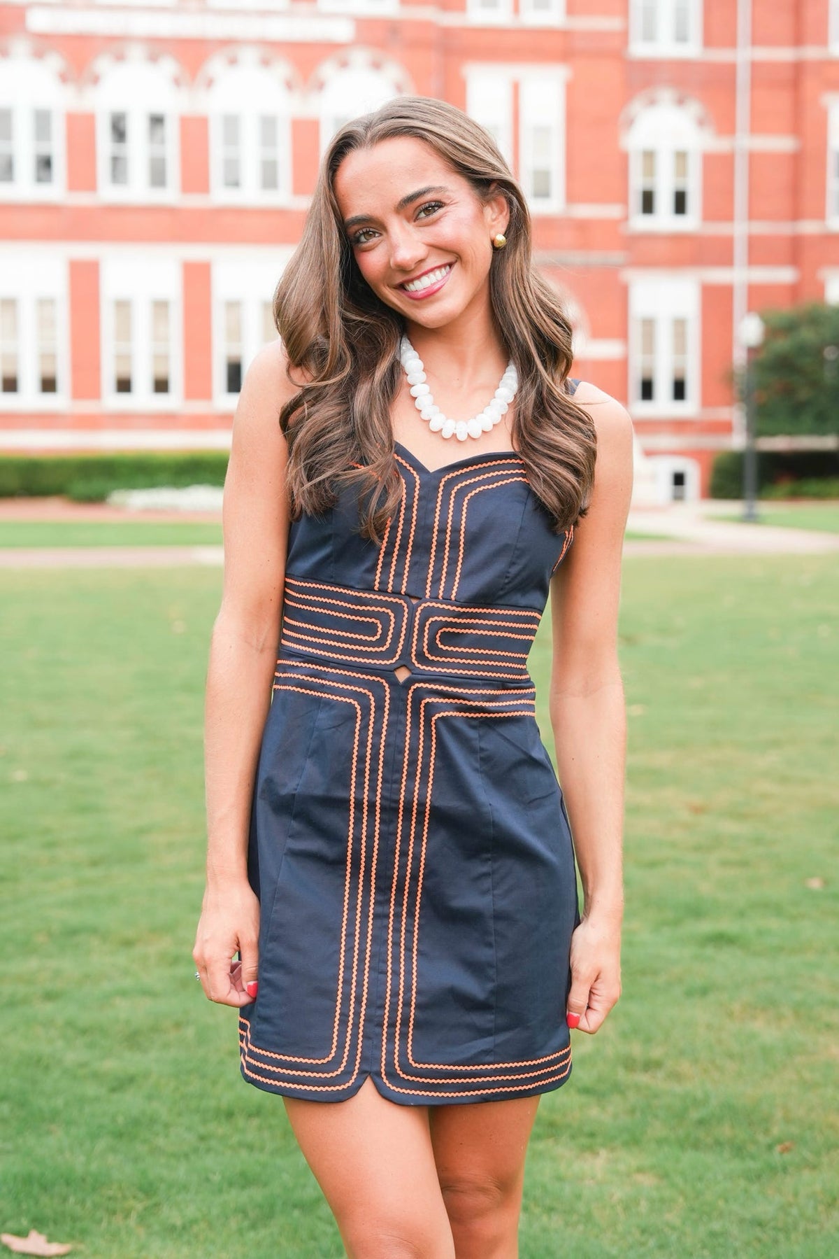 A woman wearing a sleeveless navy blue mini dress with orange geometric stripe patterns, standing on a green lawn with a red brick building in the background.