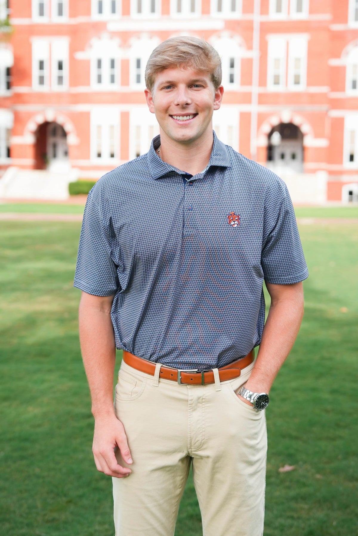 A smiling young man standing outdoors on a green lawn in front of a large red brick building, wearing a short-sleeve blue patterned polo shirt, khaki pants, and a brown belt.