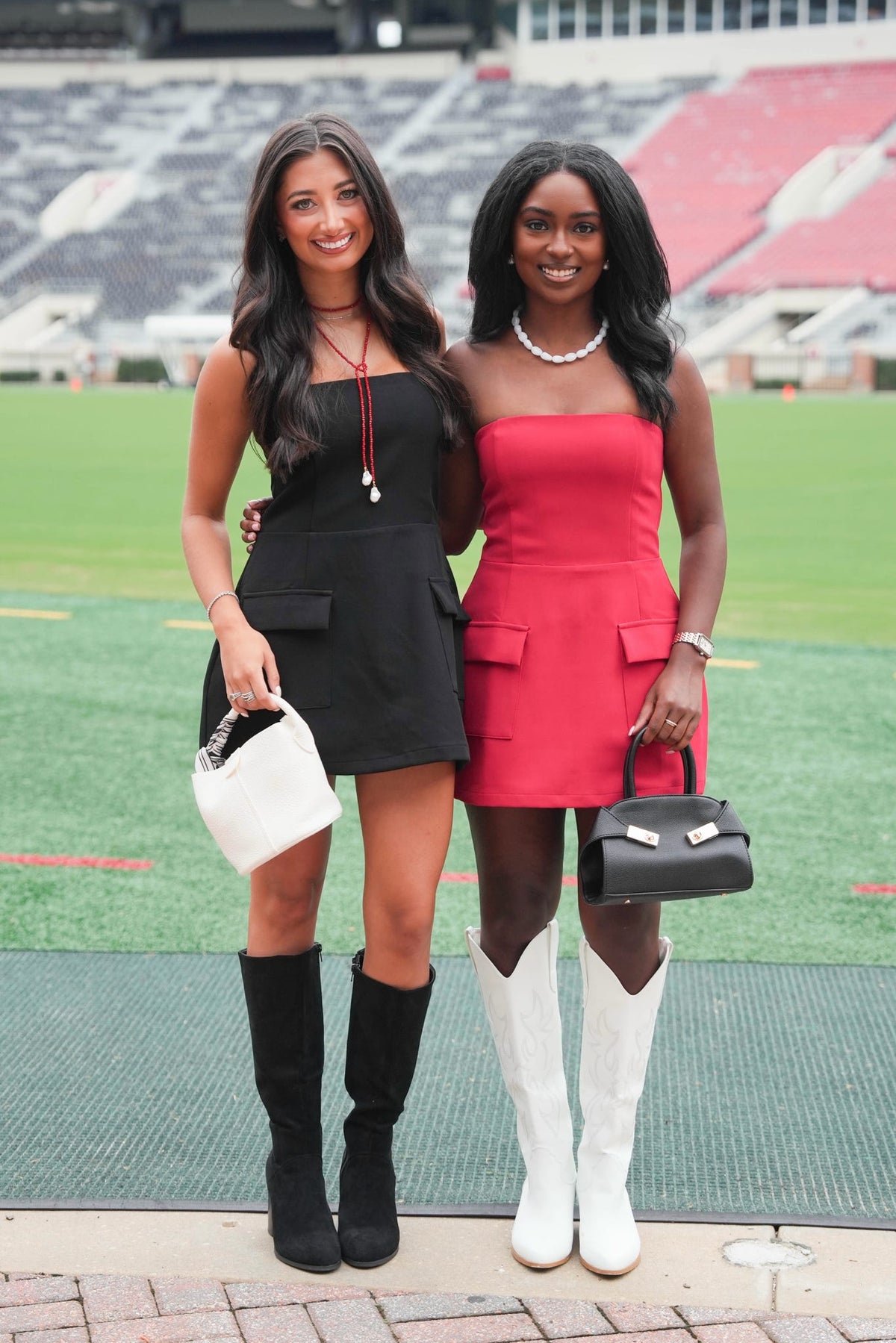 Two women posing together on a sports field in front of stadium seating. The woman on the left wears a black strapless mini dress with black knee-high boots and carries a white handbag.