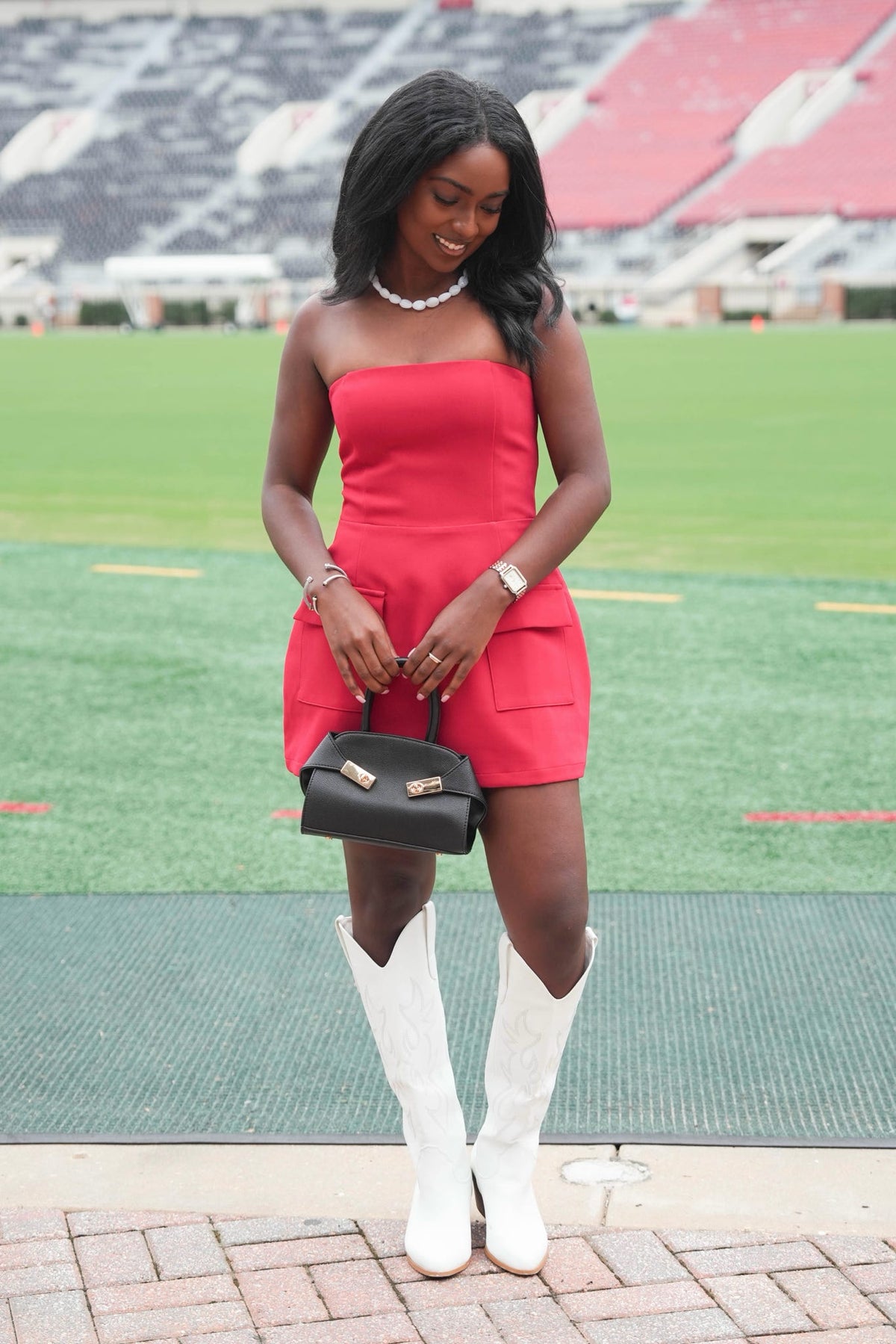 A woman stands on a brick walkway in front of a football stadium field, wearing a bright red strapless mini dress, white cowboy boots, and holding a black handbag.
