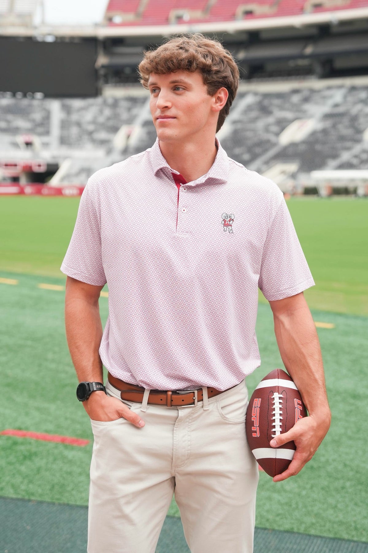 A man standing on a football field wearing a light pink polo shirt with a small embroidered logo on the left chest, paired with khaki pants and holding a football.