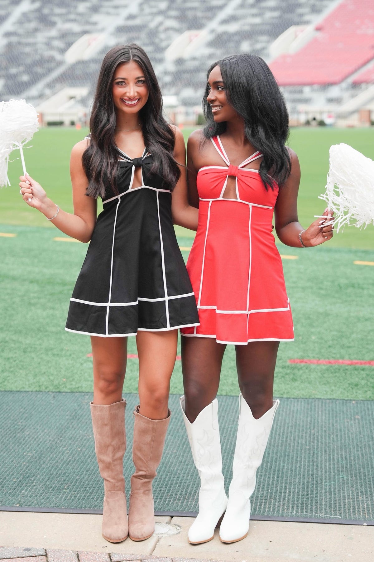 Two women modeling mini cheerleader dresses on a football field. The woman on the left wears a black dress with white piping and tan knee-high boots, holding a white pom-pom.