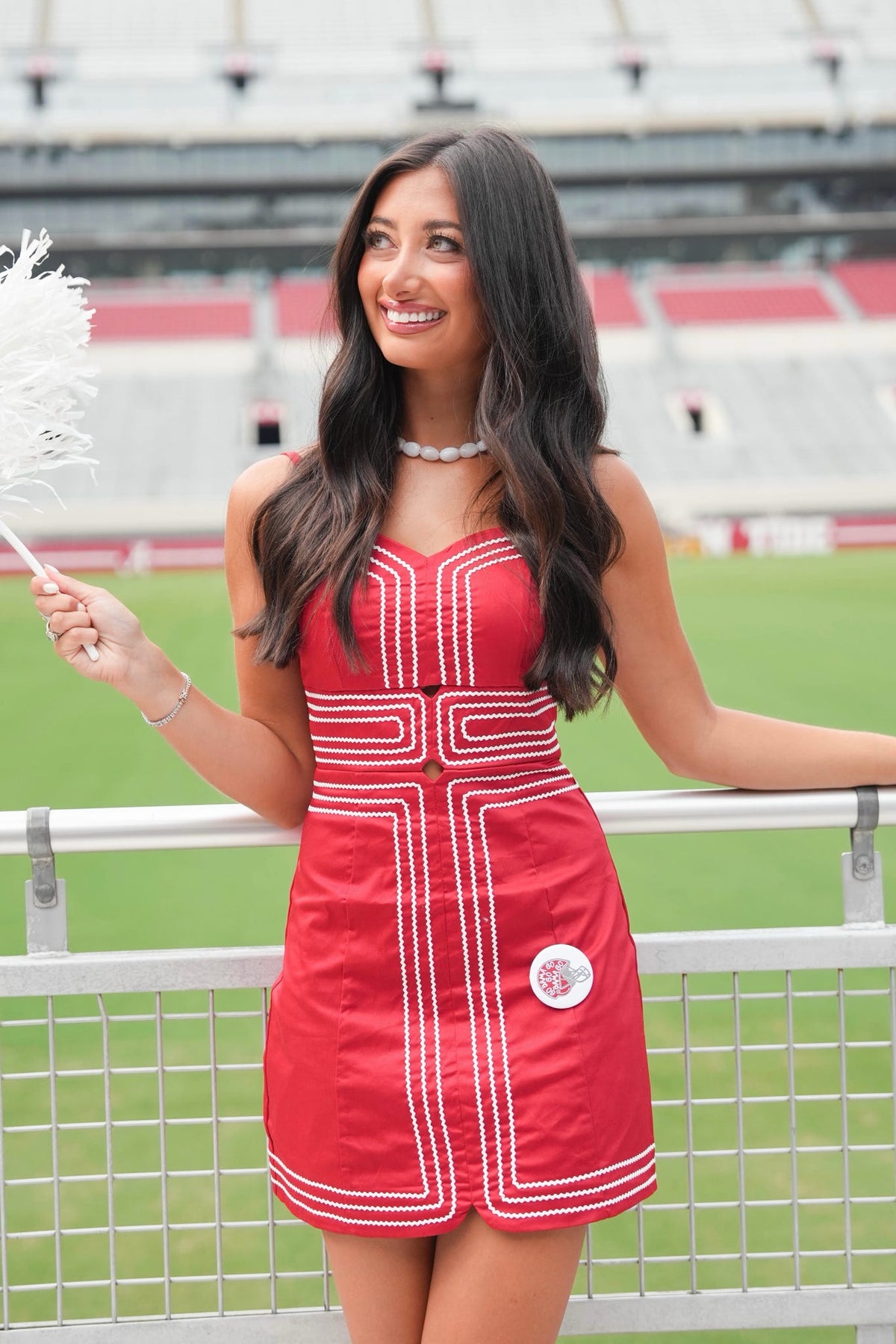A woman stands in a stadium wearing a bright red sleeveless dress with white geometric trim and a small circular patch near the hem. She is smiling and holding a white feather fan, with stadium seating and field visible in the background.